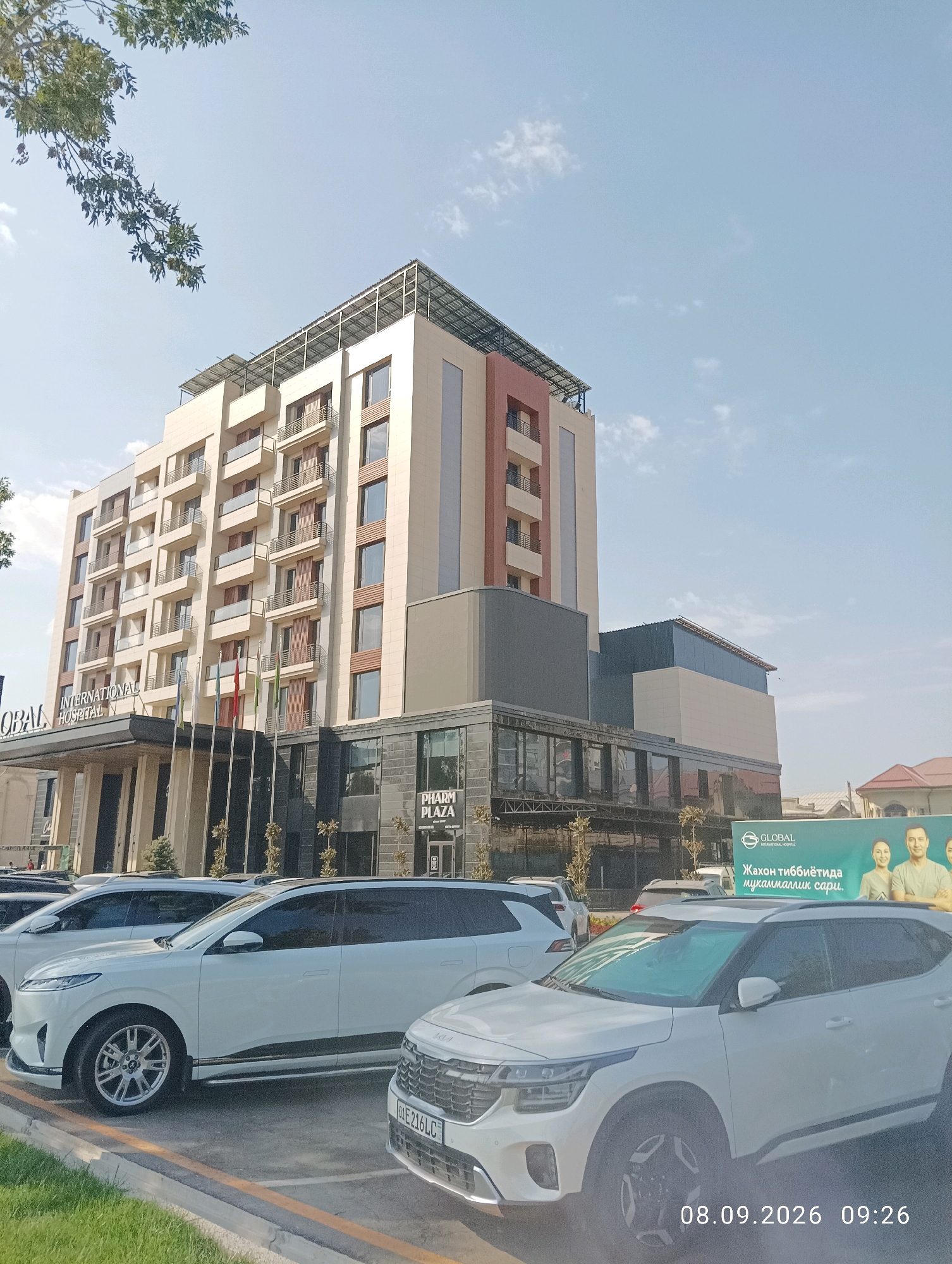 In Tashkent, Uzbekistan, a modern, multi-story building, identified as GLOBAL INTERNATIONAL HOSPITAL, stands tall against a clear blue sky. The hospital features a beige facade with brown accents and numerous balconies. A dark-colored entranceway with pillars leads to the building. In front of the hospital, a row of white SUVs is parked. A large billboard with images of medical professionals and text in Uzbek is visible to the right of the hospital. The timestamp "08.09.2026 09:26" is embedded in the lower right corner of the image.