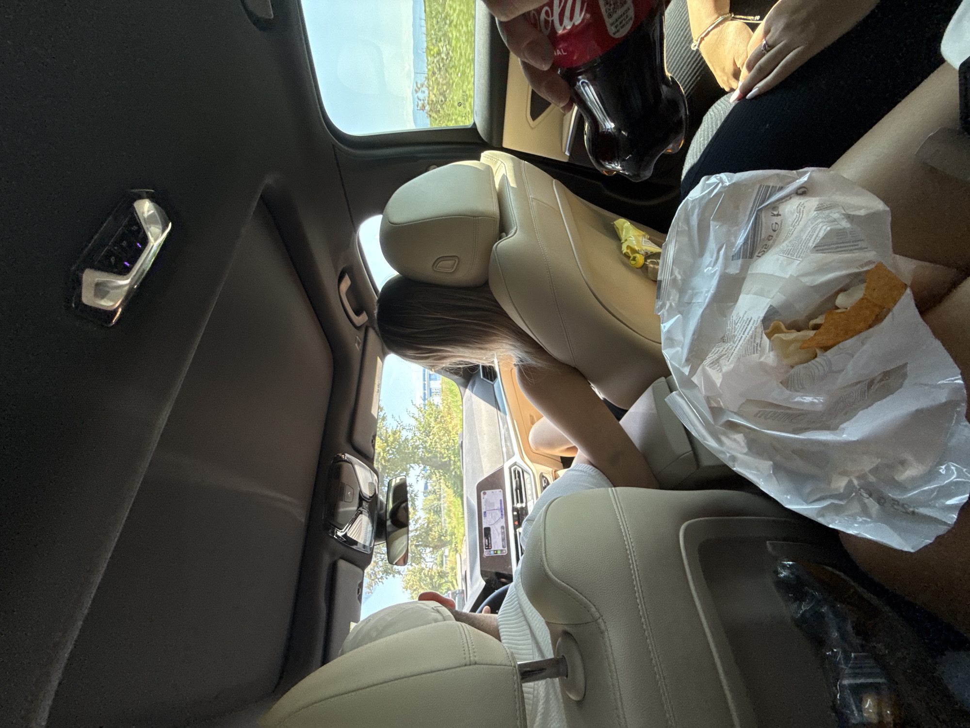 The image depicts the interior of a car, likely a luxury model given the tan leather seats and modern dashboard. The perspective is from the rear passenger seats, looking forward. A man is driving, wearing a white sweater. A woman with blonde hair is in the front passenger seat, leaning back. In the foreground, a bag of chips and a bottle of Coca-Cola are visible, with a hand reaching for the bottle. The car's sunroof is open, revealing a clear blue sky and green trees. The dashboard displays a GPS navigation system. The overall scene suggests a road trip or a casual outing. The mention of Târgu Secuiesc, Romania, is not directly supported by any visual elements in the image.