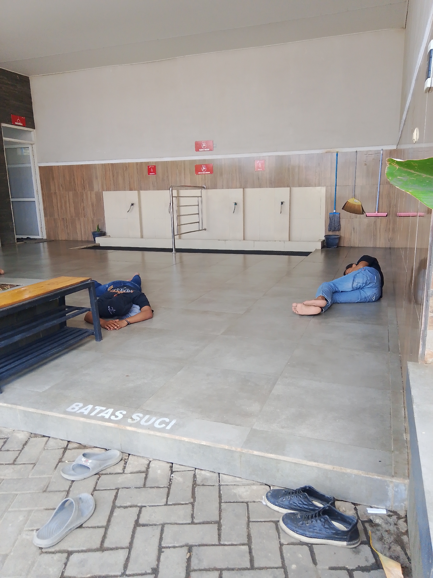 This is a wide-angle, eye-level shot taken in Gedangan, Indonesia, likely at a public facility with washing stations. Two individuals are seen sleeping on the tiled floor. The person on the left is lying face down, wearing a black t-shirt and blue jeans, with their hands clasped behind their head. They are positioned near a wooden bench with metal shelving. The person on the right is curled on their side, also wearing blue jeans and a dark t-shirt, with their back towards the camera. They are barefoot.
The environment features a row of white washing stations with faucets against a wall paneled with light brown wood-like tiles. Red signs with pictograms are mounted on the wall above the washing stations. In the background, there are cleaning supplies, including two brooms and a bucket. The floor is tiled with large grey tiles, and a marked area with the text "BATAS SUCI" is visible near the foreground. Several pairs of footwear, including flip-flops and sneakers, are scattered on the brick-patterned pavement in front of the tiled area. The overall scene is brightly lit by natural sunlight.