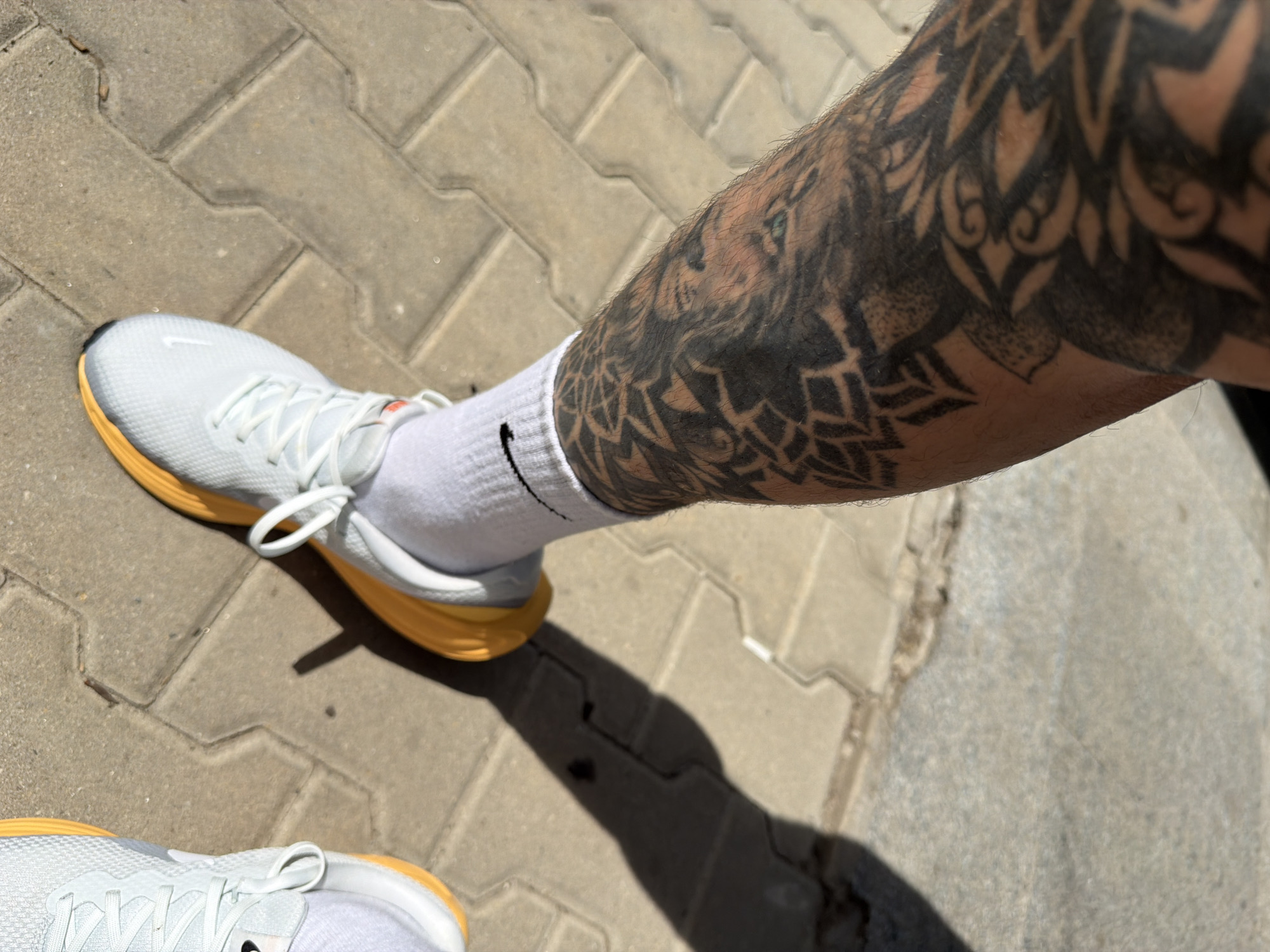The image shows a person standing on a paved surface. The person is wearing white athletic shoes with yellow soles and white socks with a black Nike swoosh. The person's leg is heavily tattooed with a black and grey design that includes what appears to be a lion's head. The pavement is made of interlocking blocks of a light grey color. Another shoe, similar in style and color to the one in focus, is partially visible at the bottom left corner of the frame.