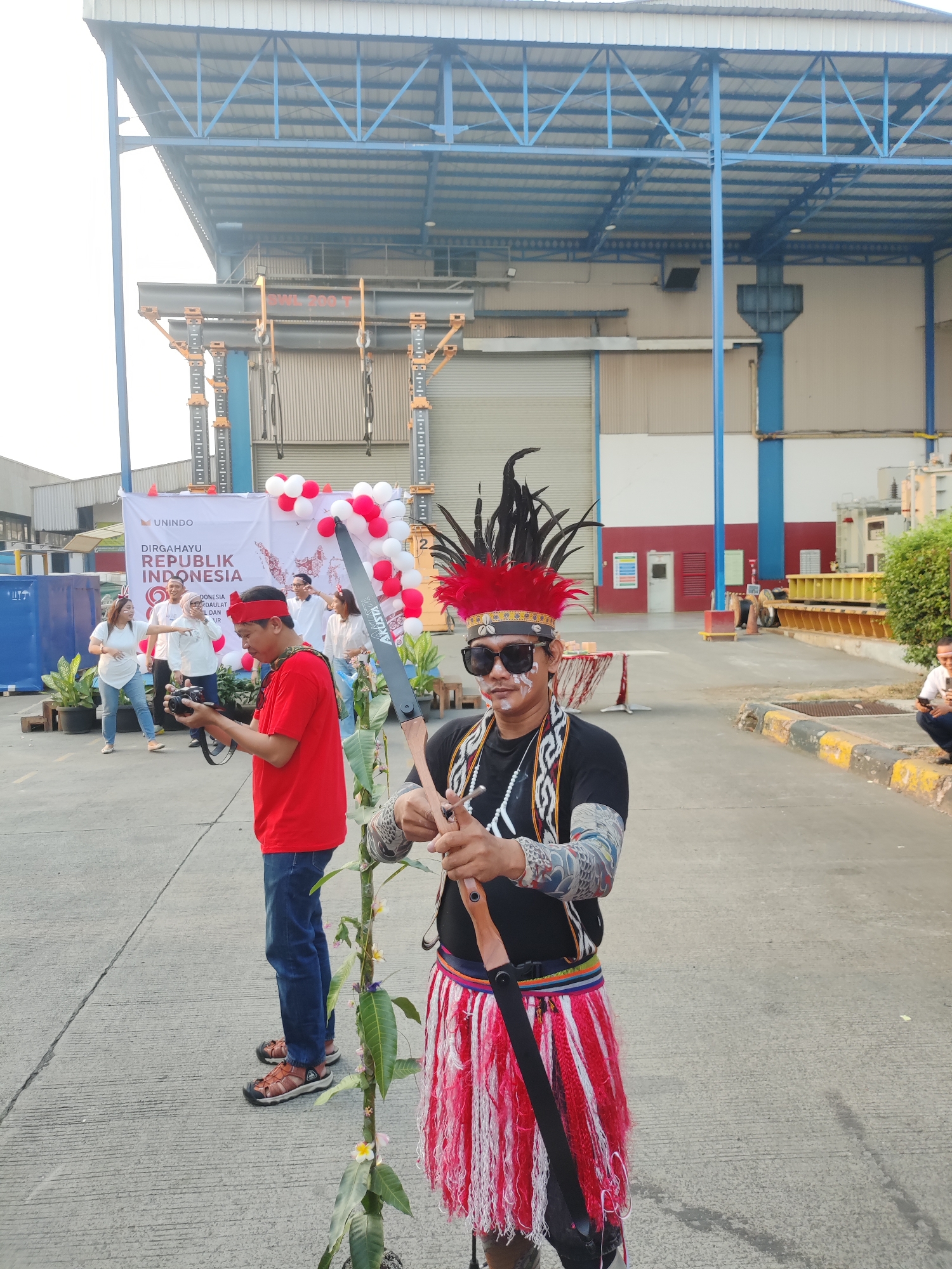 In East Jakarta, Indonesia, an outdoor event is taking place, likely a celebration given the decorations and attire. A man in traditional attire, including a feathered headdress, face paint, sunglasses, and a fringed skirt, stands in the foreground holding a bow and arrow. He appears to be posing or demonstrating the archery equipment. To his left, a man in a red shirt and blue jeans is holding a camera, possibly taking a photo of the man in traditional dress or the surrounding event. Behind them, a banner with "DIRGAHAYU REPUBLIK INDONESIA" is displayed, adorned with red and white balloons. Several other individuals are visible in the background, some interacting and others observing. The setting appears to be an industrial or open-air facility, with large buildings and a paved ground. The lighting suggests it is daytime.