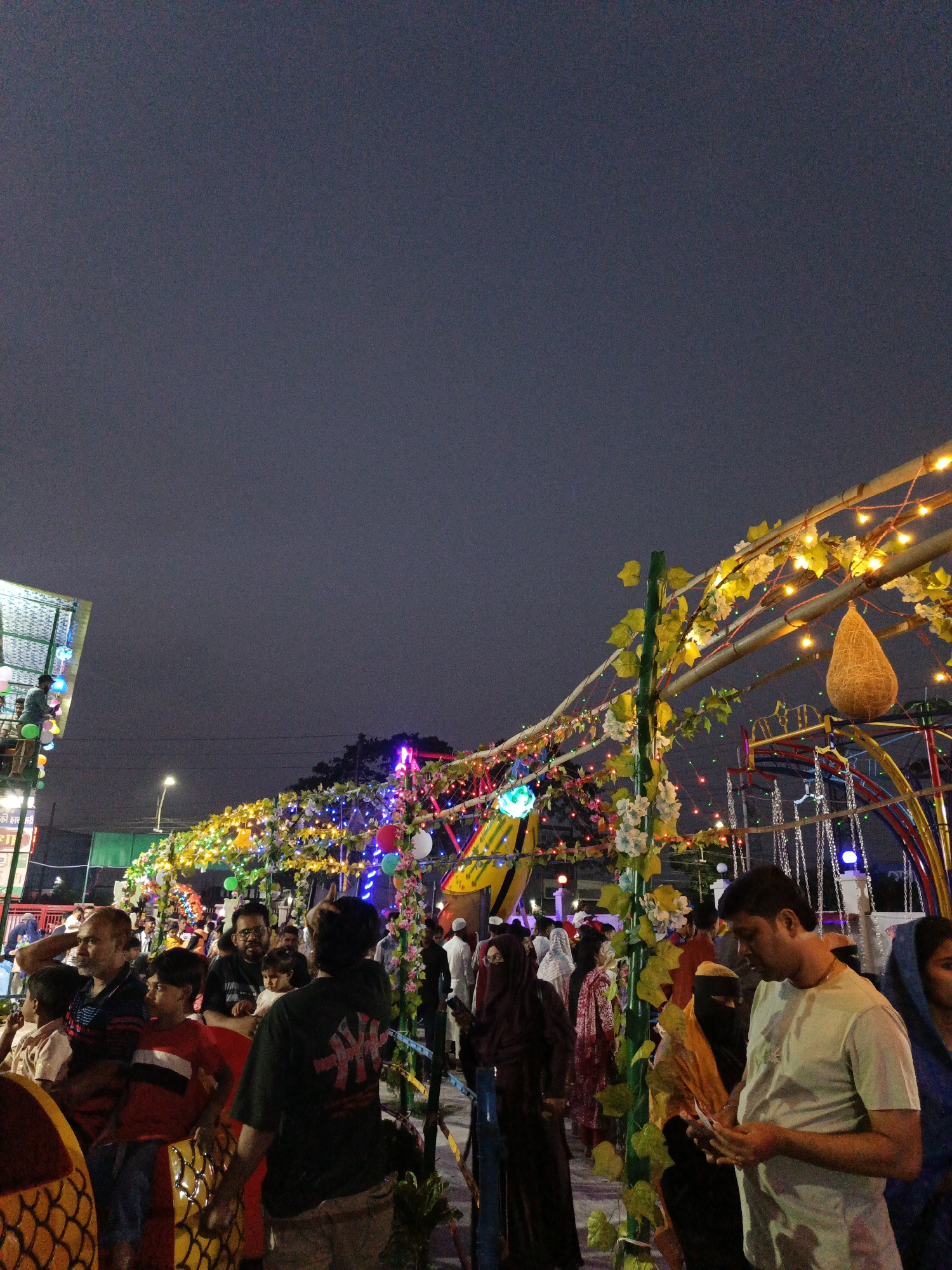 The image captures a crowded scene in Dhaka, Bangladesh, likely during a festival or event. Numerous people are gathered, including men, women, and children, some wearing traditional Islamic attire such as hijabs. The area is decorated with lights, flowers, and colorful ornaments strung along a bamboo-like structure. In the background, there are amusement park rides, suggesting a fair or carnival setting. The sky is a dusky gray, indicating it's either evening or an overcast day. A man in the foreground is looking at his phone, while others interact with their families and the surroundings.
