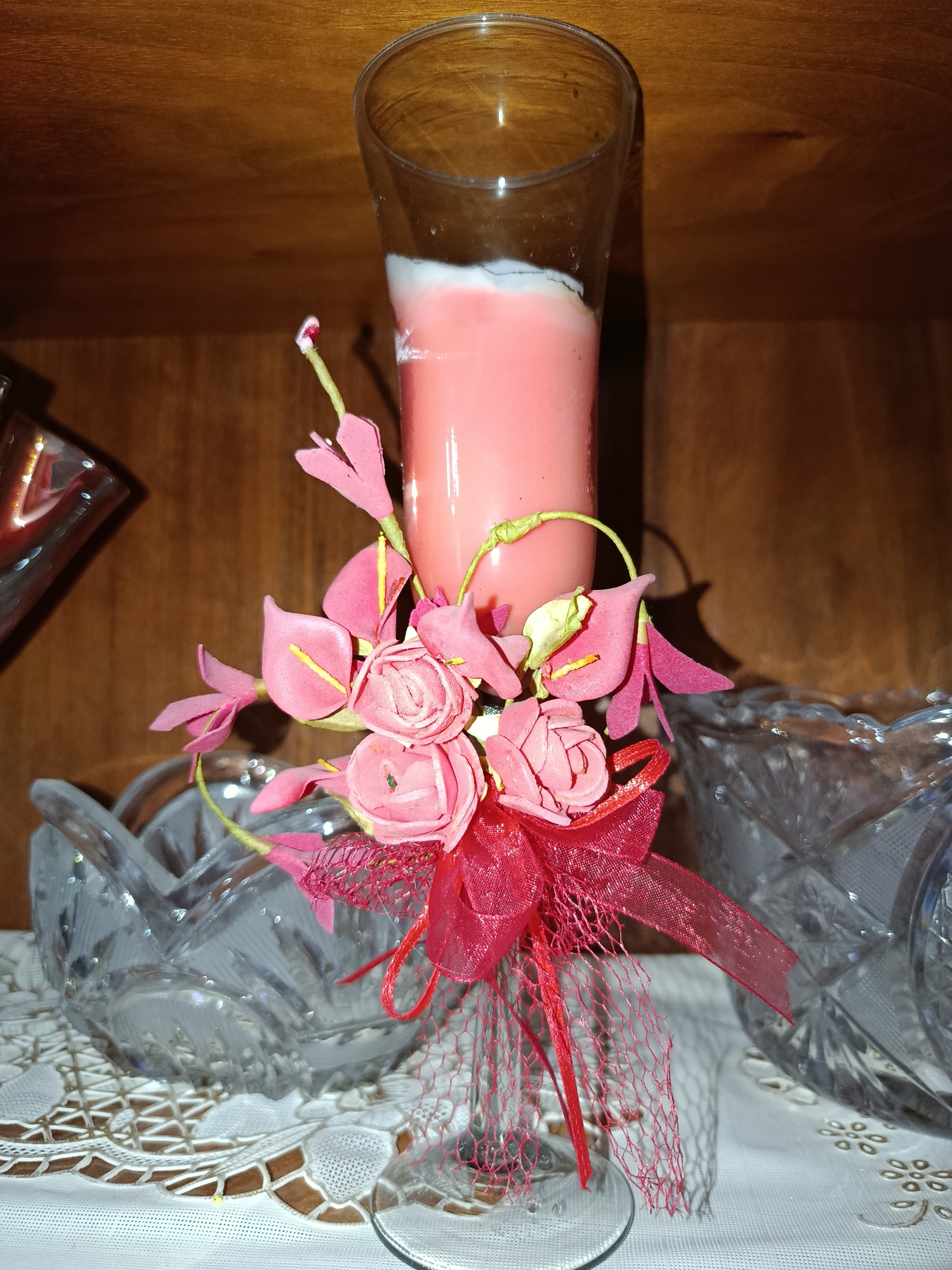 The image depicts a still life composition on a wooden shelf. In the center, a tall, clear glass contains a pink candle with a white top layer. The candle is adorned with a cluster of artificial pink flowers and a red ribbon bow with red netting. To the left and right of the candle, two ornate crystal bowls are partially visible. The background is a wooden surface, suggesting the items are displayed on a shelf or in a cabinet. The lighting is warm, highlighting the textures of the glass, crystal, and wood. No people are present in the image, and no specific actions or interactions are taking place. The scene appears to be an indoor decorative arrangement.
