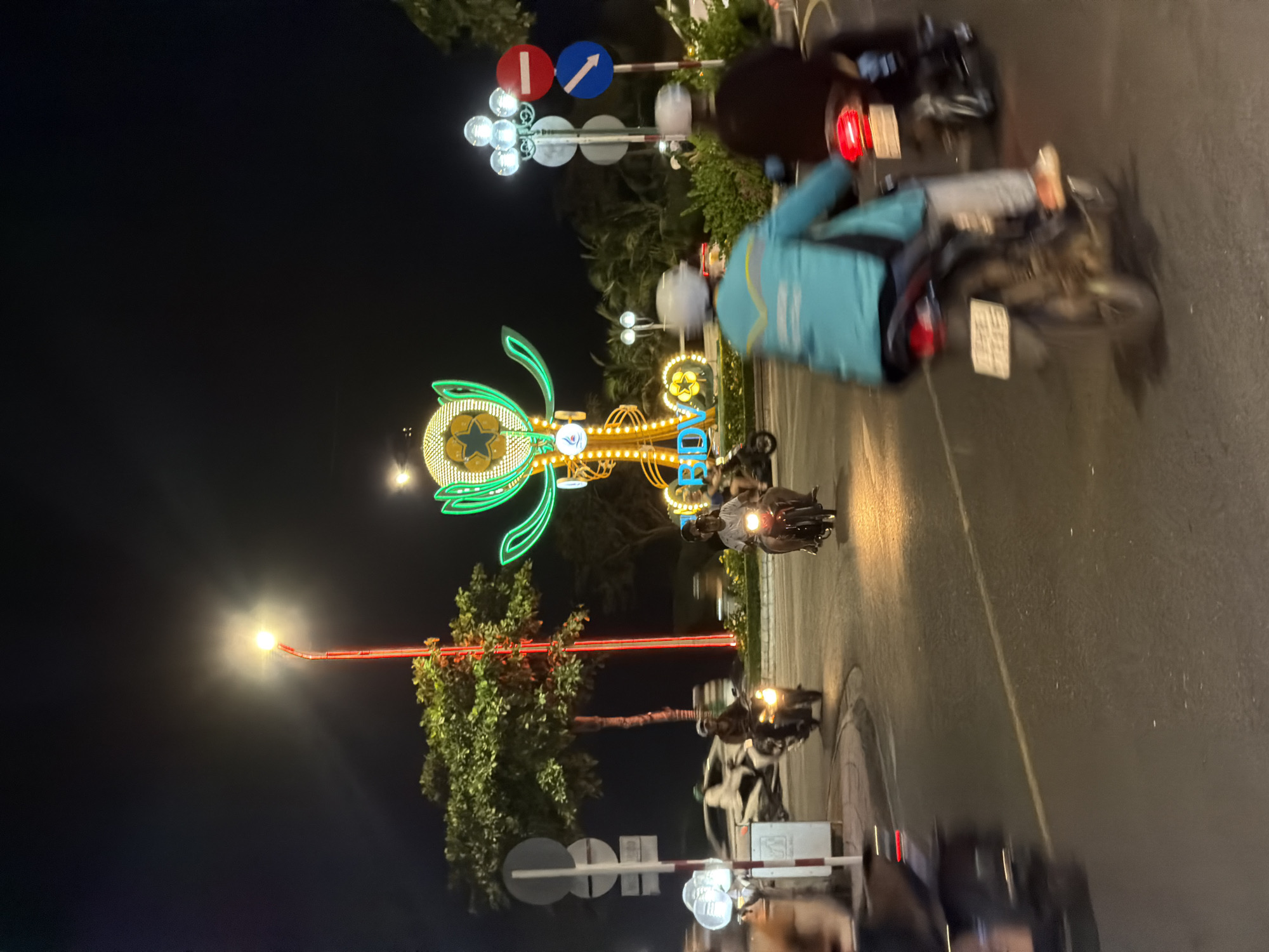 The image displays a nighttime urban street scene in Nha Trang, Vietnam. A large, brightly illuminated decorative structure dominates the central background, featuring a golden, spiral-lit column topped with a green, abstract, cup-like design containing a star motif. The blue letters "BIDV" are lit near its base.
The foreground and mid-ground are occupied by multiple motorcycles in various states of motion. Two motorcycles in the immediate foreground are blurred, indicating movement away from the viewer; one rider wears a teal jacket, and the other a dark jacket, both displaying illuminated red tail lights. Further back, two other motorcyclists, wearing helmets, are visible with active headlights. A white car, heavily blurred, is partially visible on the far left behind a signpost.
The roadway is asphalt with discernible white lane markings. A raised central island, planted with green foliage, hosts the primary decorative structure. Street lighting includes a tall red-poled lamp with a bright white light on the left and a multi-spherical white lamppost on the right. Road signs are present on the right, consisting of a red circular "no entry" sign and a blue circular directional arrow sign. Trees with green foliage line the street and the central island, illuminated by ambient light. The scene is lit by various artificial sources against a dark night sky.