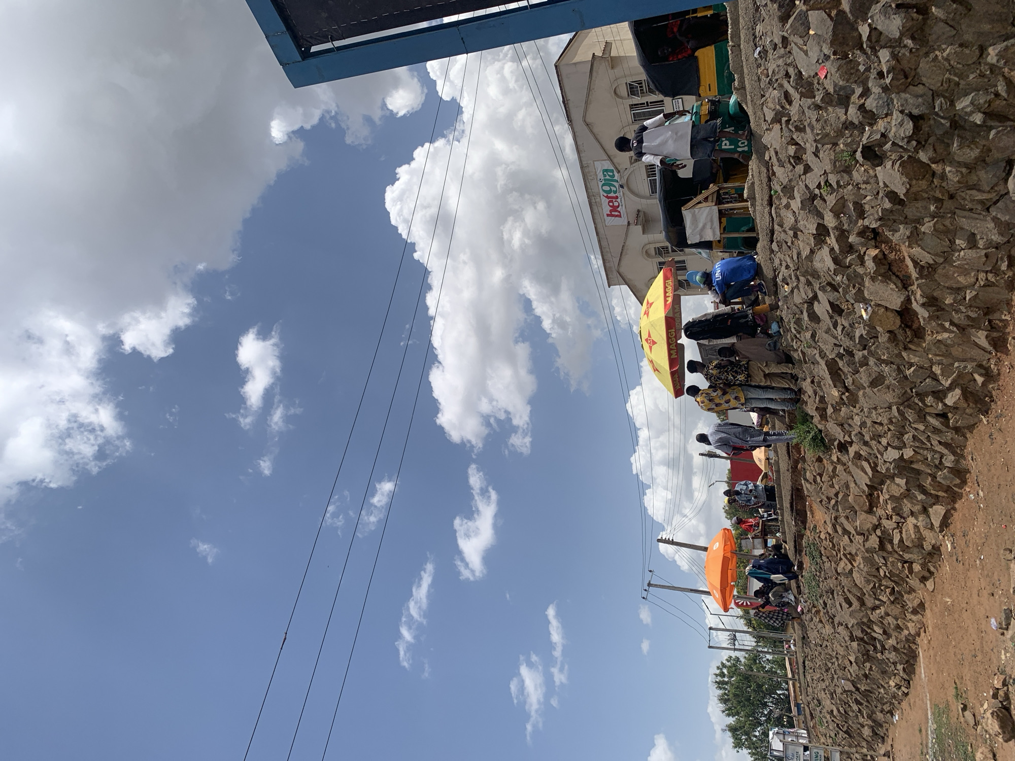 The image, initially presented with a 90-degree rotation, captures a dynamic daytime street scene in Jos, Nigeria. A bright blue sky filled with large, fluffy white cumulus clouds dominates the upper portion, with power lines stretching across it.
In the mid-ground, a multi-story building with light-colored architecture stands, featuring a prominent red and white "Bet9ja" sign on its facade. Below this, the scene unfolds into a bustling roadside environment. A rugged, rocky embankment or retaining wall runs diagonally across the frame, separating the street from the immediate foreground where people are gathered.
Numerous individuals populate the scene, appearing to be engaged in various activities typical of a street market or public gathering. Many are clustered under colorful market umbrellas for shade. A distinctive yellow umbrella prominently displays the "Maggi" brand logo, and an orange umbrella is also visible further down the street. People are dressed casually; one person wears a blue shirt with "UNITY" clearly printed on it.
A green and yellow vehicle, possibly a tricycle taxi or small bus, is partially visible near the building, bearing the text "PA 10..." on its side. Makeshift stalls or display areas are present, suggesting commercial activity. Utility poles and additional power lines are visible towards the right side of the frame, further establishing the urban setting. The overall impression is that of a lively and active community space under sunny conditions, with a mix of commerce and daily life unfolding.