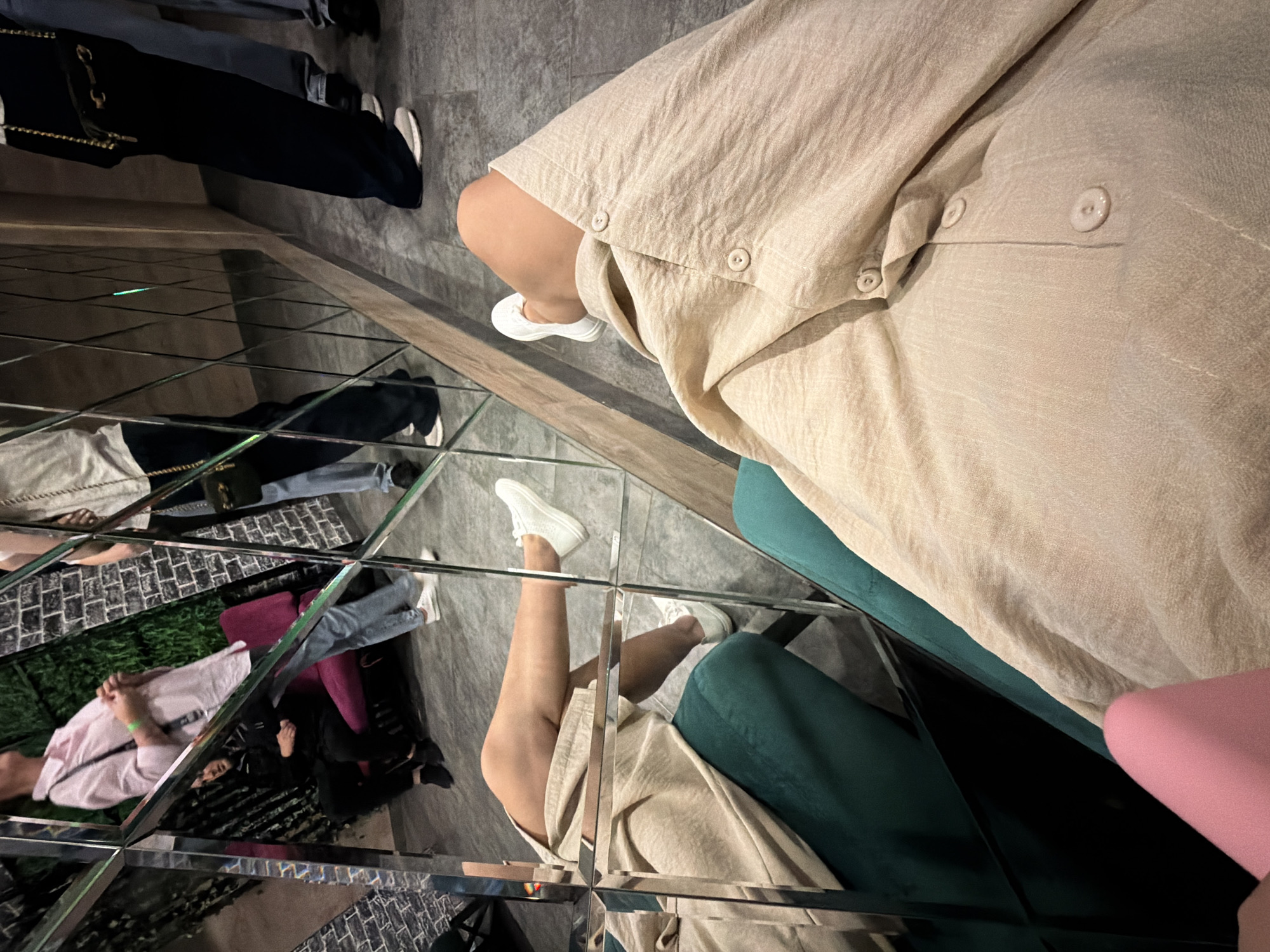 The image shows a first-person perspective of a person sitting in a room with mirrored walls. The person is wearing a light-colored linen dress and white sneakers. In the reflection of the mirror, other people are visible, including a person in a pink shirt and blue jeans, and another person in a black jacket and black pants. There are also some chairs visible in the reflection, one in pink and one in dark green. The floor is tiled. The environment appears to be indoors, possibly a lounge or club, given the decor. There is no information in the image to suggest a specific location in the city of Chaikovsky, Russia.