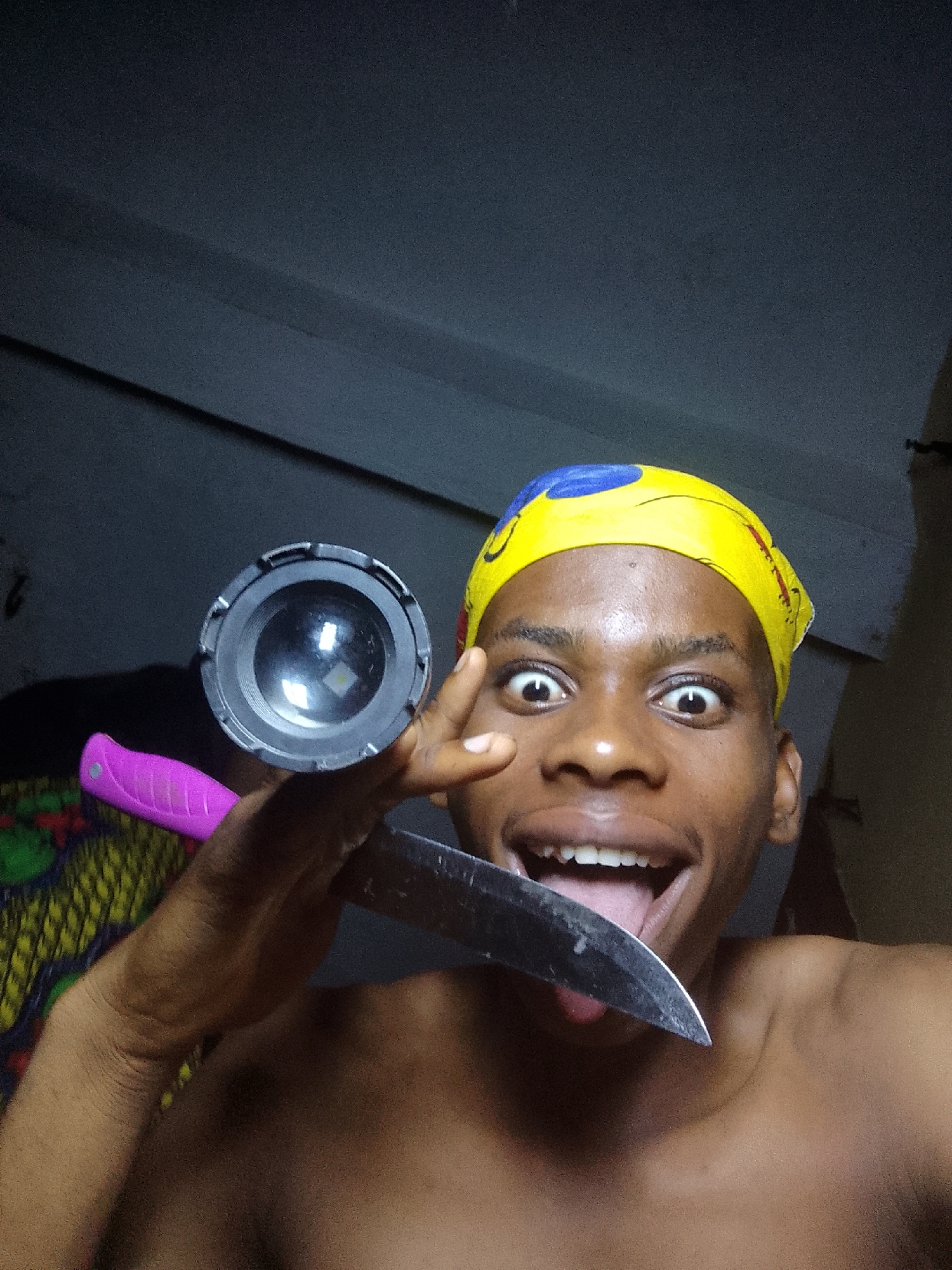 A shirtless young adult male, likely of African descent, is seen in a dimly lit indoor location. He is wearing a yellow bandana with blue and red designs on his head. He has his eyes wide open and tongue protruding from his open mouth. He is holding a flashlight with a pink handle in one hand, and a kitchen knife with the blade near his tongue in the other hand. The location is presumed to be in Nkpor, Nigeria, based on the prompt, but this cannot be verified from the image alone. The background shows a dark wall or ceiling and a partially visible patterned fabric on the left. The scene appears staged.