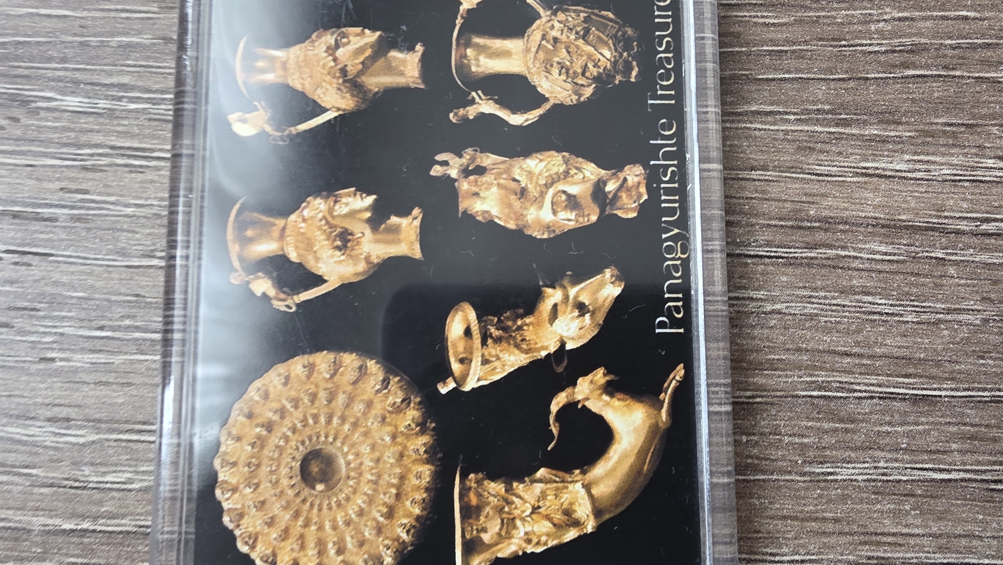 The image shows a framed card depicting the Panagyurishte Treasure, a Thracian gold hoard discovered in Panagyurishte, Bulgaria. The card features several gold artifacts against a black background, including rhytons and a phiale. The text "Panagyurishte Treasure" is visible on the right side of the card. The card appears to be in a clear plastic frame and is resting on a wooden surface. The city of Blagoevgrad is not directly represented, but as part of Bulgaria, the Panagyurishte Treasure is considered part of Bulgaria's cultural heritage and may be exhibited in Blagoevgrad.