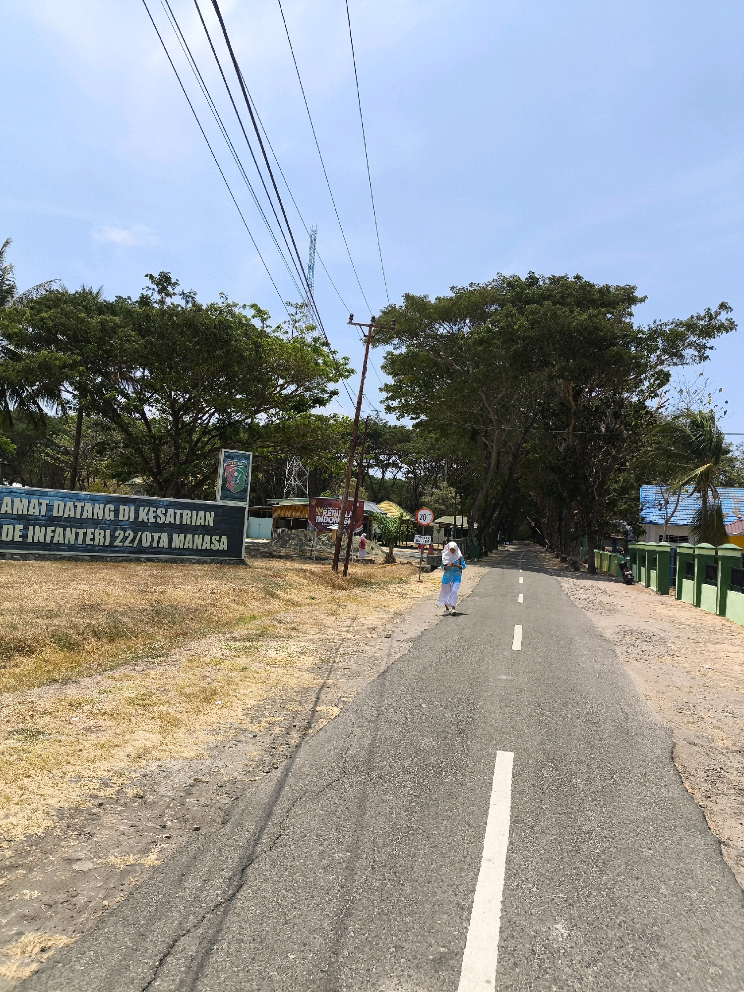 The image depicts an outdoor scene on a sunny day. In the foreground, a paved road with white lane markings runs through a tree-lined avenue. A single individual wearing a light blue hijab and matching garment walks away from the camera on the right side of the road. To the left of the road, there is a dry, grassy area and a long, dark sign with white text that reads "SELAMAT DATANG DI KESATRIAN BRIGADE INFANTERI 22/OTA MANASA." Behind the sign, a large, lush green tree dominates the landscape, with other trees and foliage visible in the background. Several utility poles and overhead wires are present, stretching across the sky. Further down the road, on the right side, a green fence runs along the edge of the property, with some buildings visible behind it. A speed limit sign indicating "20" is also visible near the entrance. The overall environment suggests a rural or semi-rural setting, possibly near a military installation, in Indonesia.