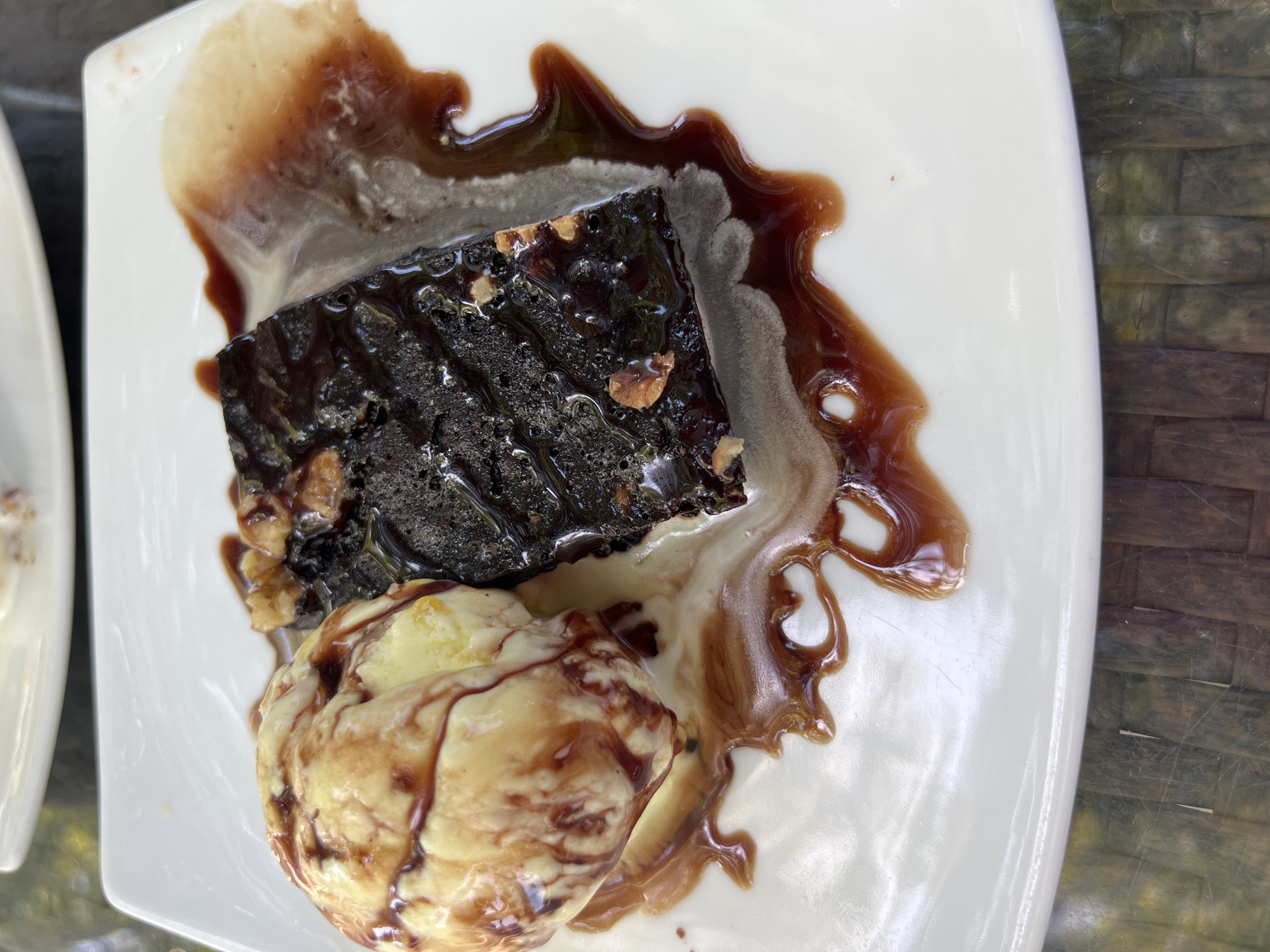 A dessert is served on a white plate. On the left is a scoop of vanilla ice cream drizzled with chocolate sauce. To the right of the ice cream is a rectangular brownie, also drizzled with chocolate sauce and topped with nuts. The ice cream and brownie are beginning to melt and blend together on the plate, with chocolate sauce pooling around them. The plate sits on a dark, textured surface, possibly a table. The scene is viewed from directly above. The location of this dessert cannot be determined from the image alone.