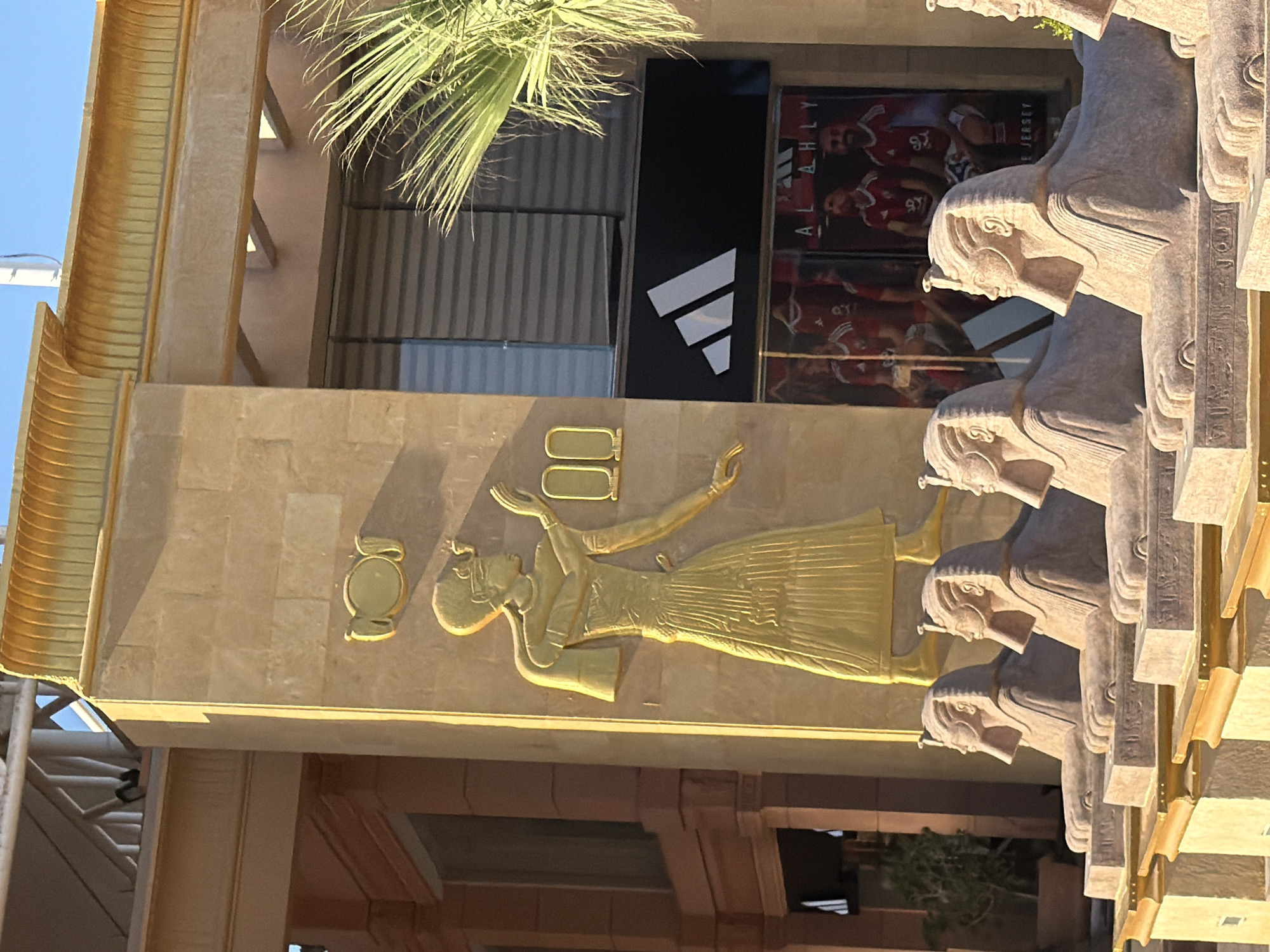 The image shows an outdoor scene in Sharm El Sheikh, Egypt, featuring a building with Egyptian-themed architecture. In the foreground, a row of four sphinx statues with stone bases is visible. The sphinxes are facing left and have detailed carvings on their bases. Behind the sphinxes, a wall of a building is decorated with a large, golden, relief sculpture of an ancient Egyptian figure wearing a headdress and a long, pleated skirt. The figure's arms are raised, and there are two small, oval gold reliefs to its right. Above the relief, the building has a decorative golden cornice. To the right of the relief sculpture, there is a storefront with large windows displaying merchandise. A black sign with a white Adidas logo is mounted above the windows, and advertisements featuring soccer players in red jerseys are visible inside. A palm frond extends into the upper right corner of the frame. The lighting suggests it is daytime, with sunlight casting shadows.