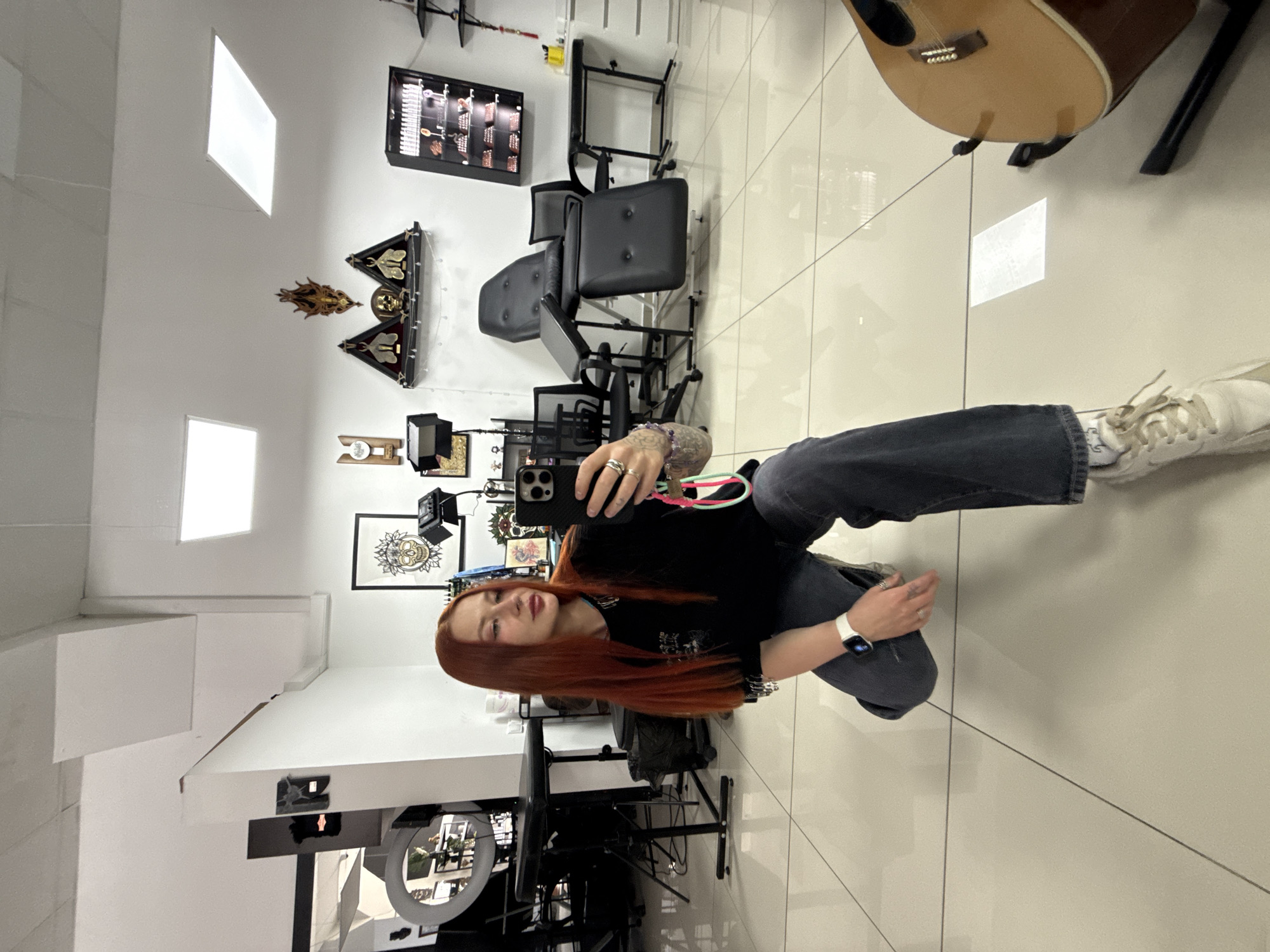 A young woman is kneeling in what appears to be a tattoo parlor. She is holding a smartphone and taking a selfie. The room contains multiple chairs, tattoo equipment, and framed artwork on the walls. A guitar leans against a wall in the background. There is no indication of the location being Rostov-on-Don, Russia.