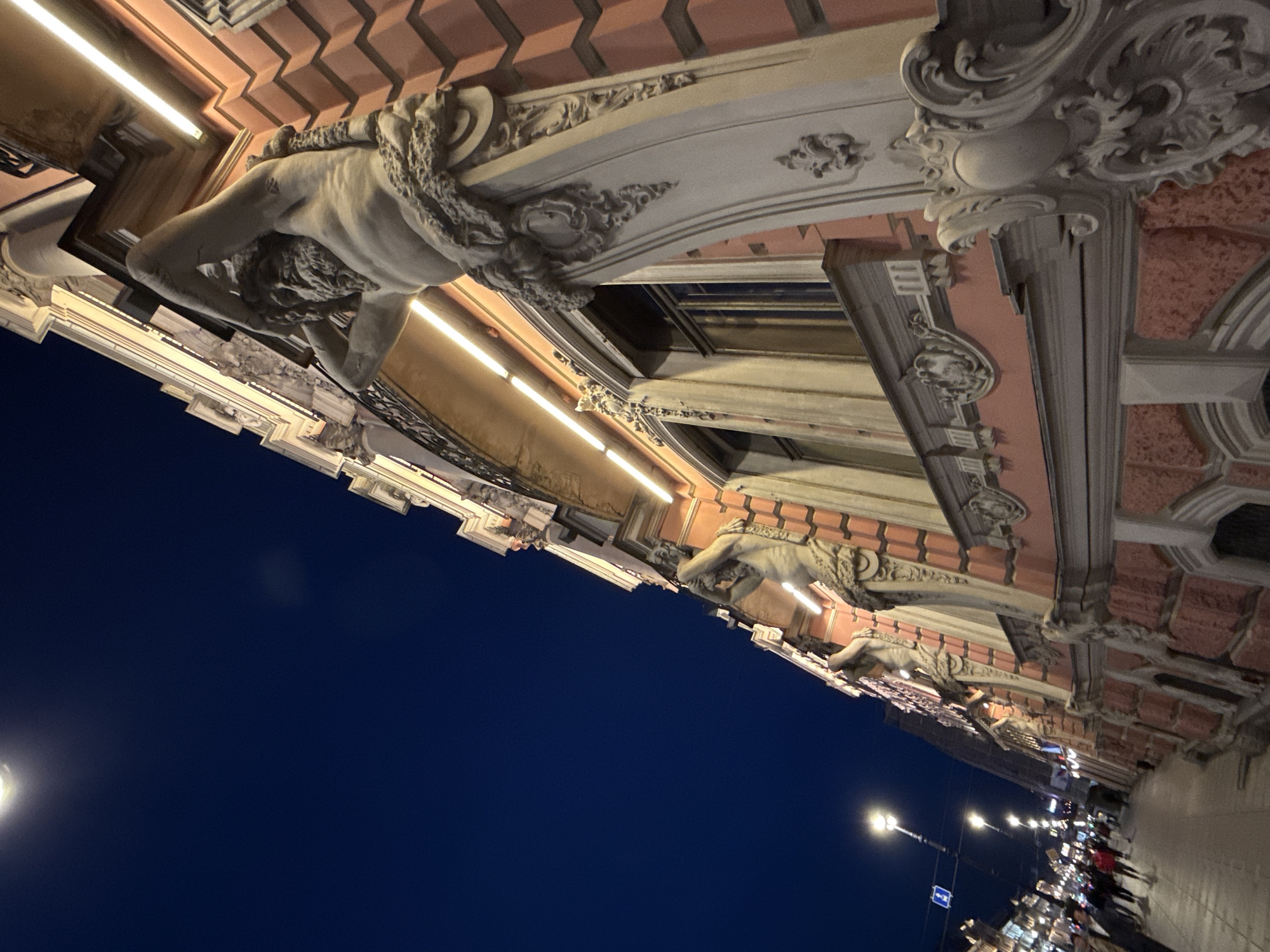 The image captures a low-angle view of the facade of a building at night, likely in Saint Petersburg, Russia, given the architectural style and typical urban setting. The building features ornate decorative elements, including sculptures of male figures (atlantes) holding up the structure, and intricate relief work. The facade is painted a salmon pink color, with cream-colored window frames and a white decorative cornice. Linear lights are installed beneath the upper floor of the building, illuminating the facade and highlighting the architectural details. The sky is a deep, dark blue, indicating it is nighttime. In the background, a street is visible with streetlights, lampposts, and pedestrians walking along a sidewalk. The perspective creates a dramatic and imposing feel, emphasizing the grandeur of the architecture.