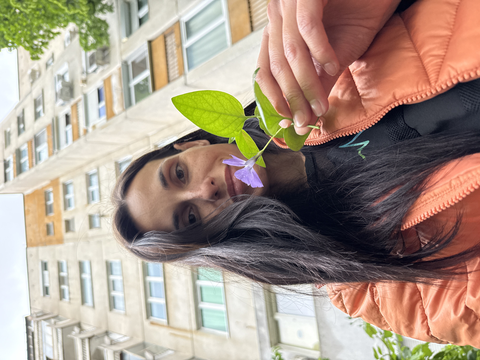 A woman with long, dark hair is holding a sprig of green leaves and a small purple flower. She is wearing an orange puffer jacket over a black shirt. The background is an urban landscape. The location is likely Pleven, Bulgaria. There is a multi-story building with numerous windows behind her.