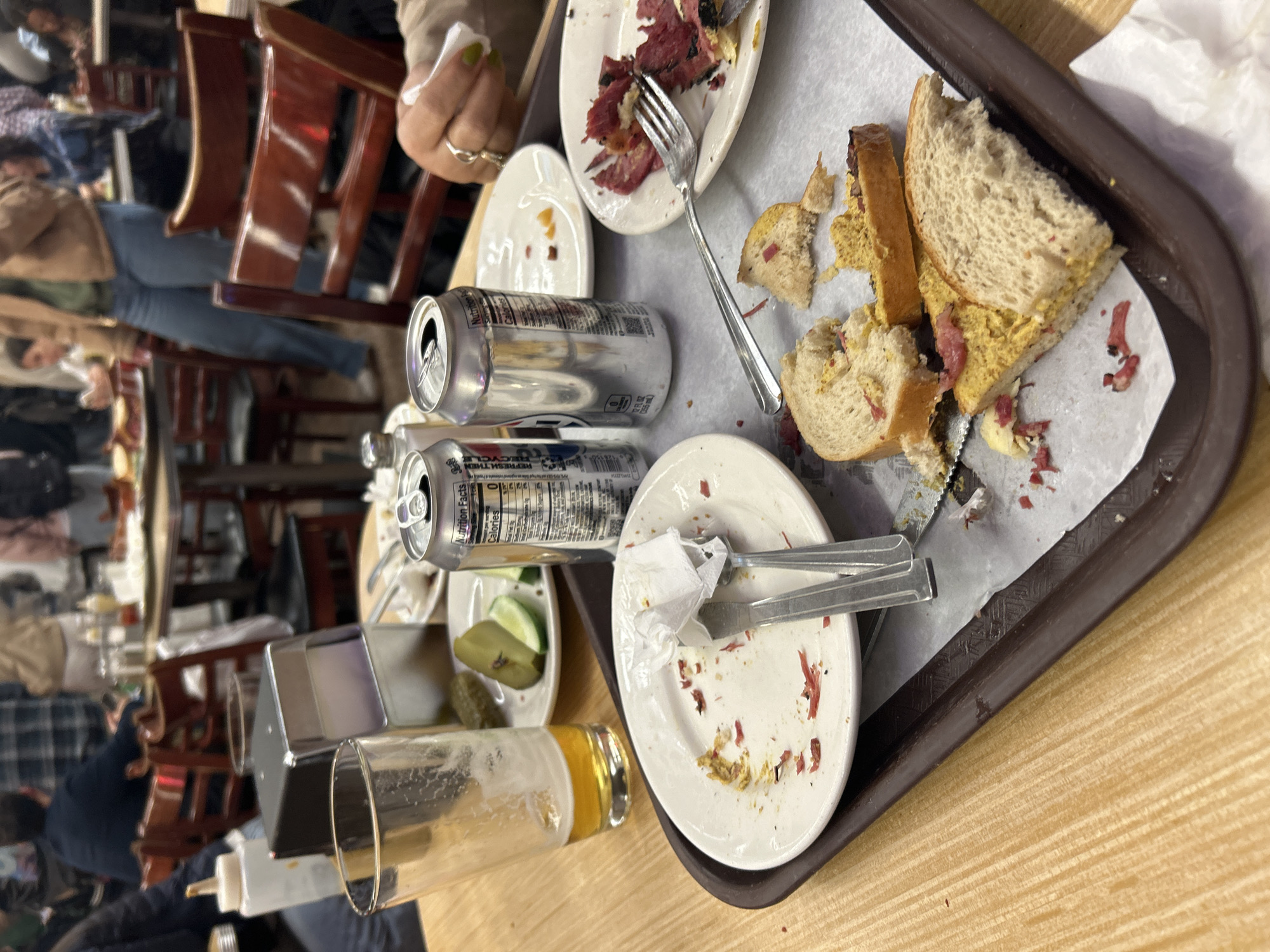 The image depicts a partially cleared table setting at Katz's Delicatessen in New York City. A brown tray contains remnants of a meal, including a plate with bits of pastrami, a fork, a disassembled sandwich with mustard, a plate with a napkin and silverware, a plate with pickle slices, a near-empty beer glass, and two open beer cans. A woman's hand with painted nails is visible at the top of the frame. The background is blurred, showing a busy interior scene with tables, chairs, and people.
