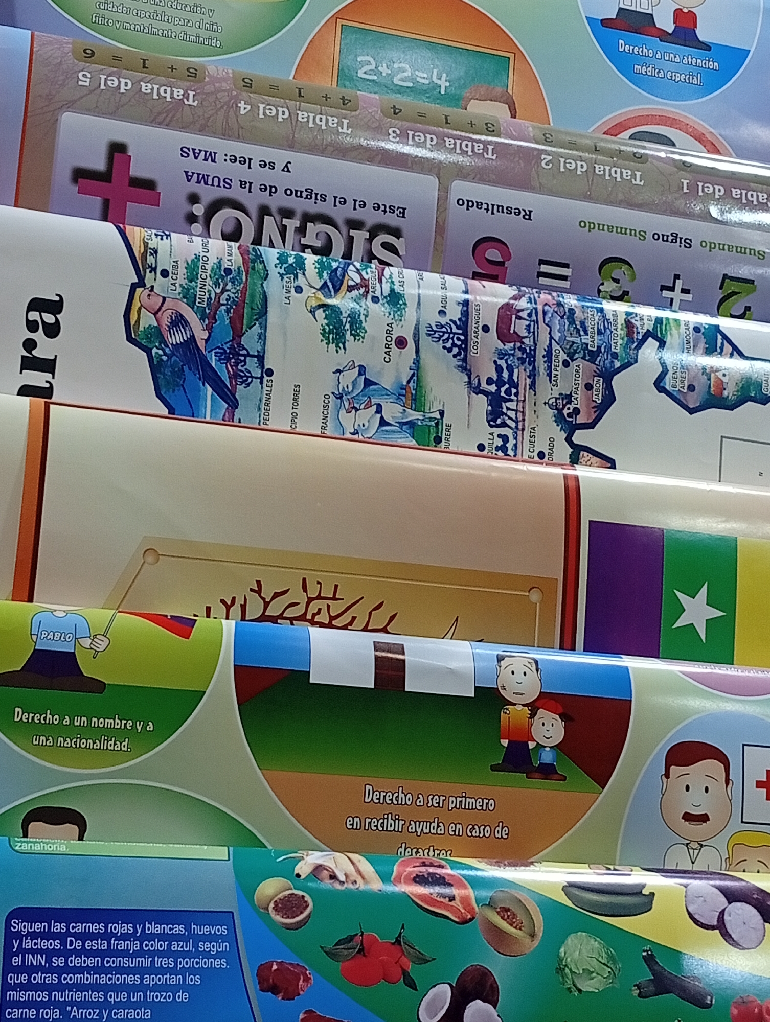 The image shows a close-up of stacked educational posters. The posters are colorful and feature various graphics and text, some of which appear to be in Spanish. One poster shows a blackboard with "2+2=4" written on it. Another poster displays a map with labels like "CARORA" and "LOS ARAGÜES," which are locations in Venezuela. There is a poster with numbers and mathematical symbols. Another poster has illustrations of people and text about "Derecho a un nombre y a una nacionalidad" (Right to a name and nationality). Further down, there are posters with illustrations of fruits and vegetables and text about healthy eating. One poster shows cartoon characters, one of whom is labeled "PABLO." The environment appears to be indoors, possibly a classroom or a storage area where educational materials are displayed or kept. There are no clear indications of the city of Juan Griego, Venezuela, within the visible details of the image, although place names on one poster suggest a connection to Venezuela. The overall impression is that of a collection of educational materials for children, likely related to math, social studies, and health.