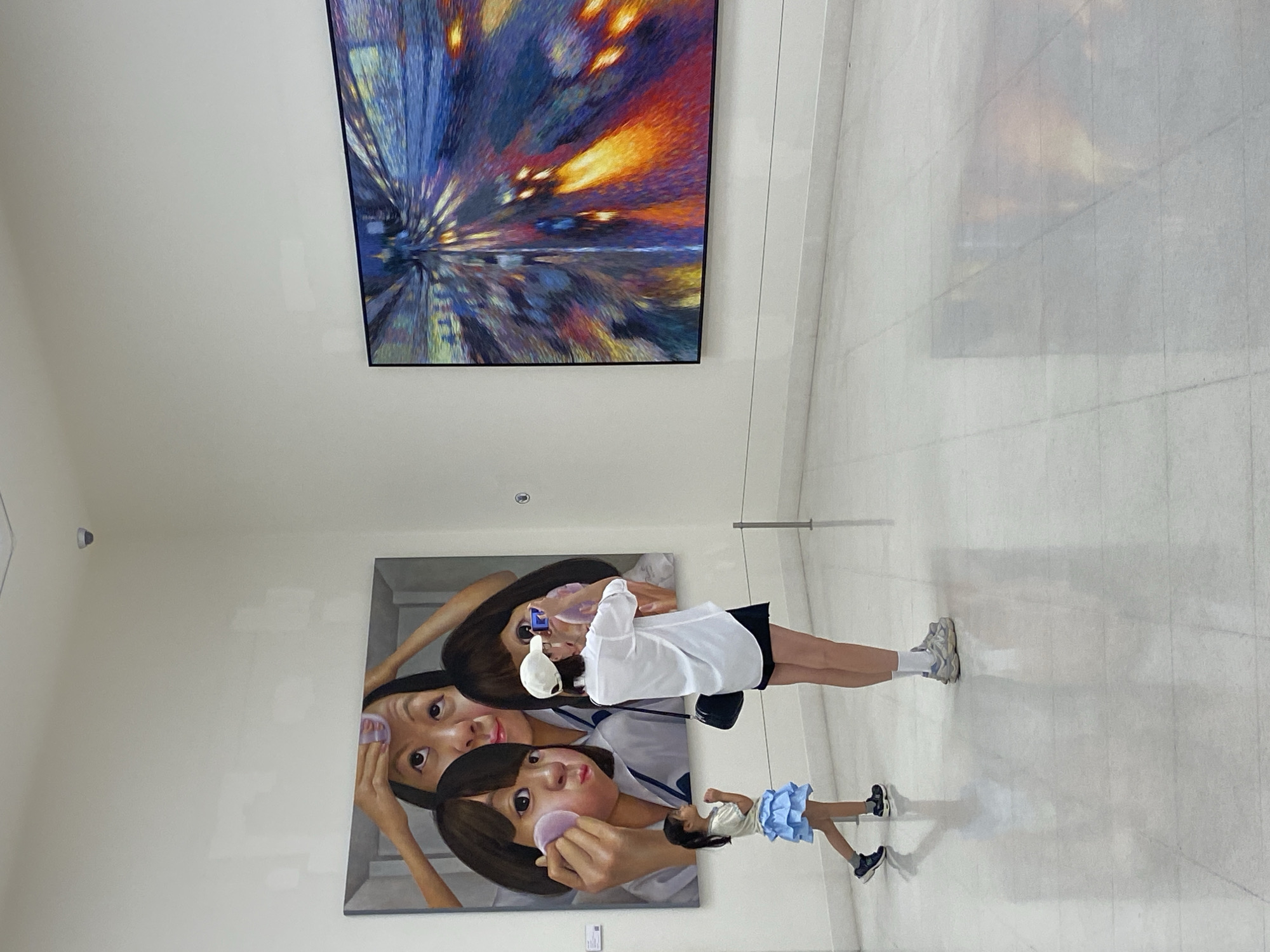 A brightly lit interior space features two artworks on a white wall. The upper artwork is an abstract piece with blue, orange, and yellow hues resembling motion or light trails. Below and to the left, another artwork depicts two women, one seemingly applying makeup to the other.
In front of the artworks, a woman wearing a white shirt, black skirt, white cap, and sneakers is taking a picture of the lower artwork with her smartphone. A young girl in a white shirt and blue ruffled skirt stands beside her. The space appears to be a modern art gallery with a reflective, light-colored floor. The image was taken in the city of Bangkok, Thailand.