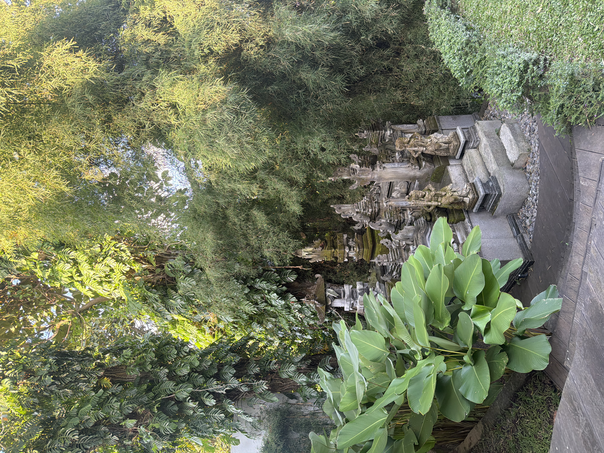 The image shows a scene in Mas, Indonesia. A stone structure, possibly a small temple or monument, is centered in the view. The structure is surrounded by lush greenery, including bamboo trees and large-leaved plants. The ground appears to be paved or covered with stones, leading to the structure. The overall environment suggests a tranquil and natural setting.