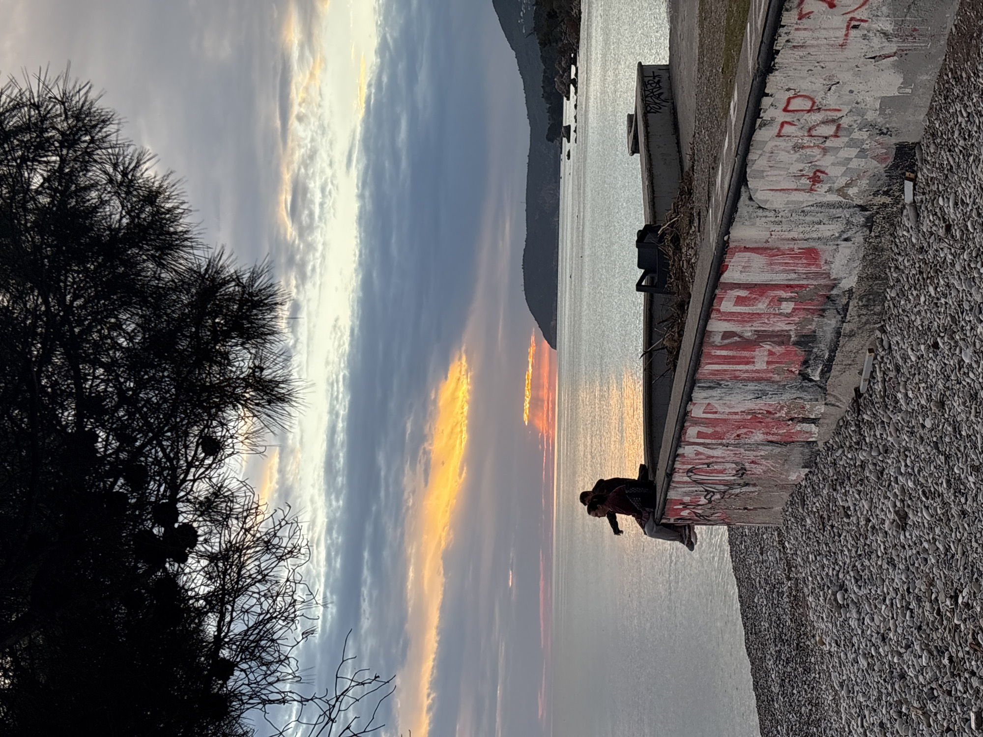 A horizontal shot of a landscape at sunset, rotated to have a tree at the left. The right side of the scene features a structure with painted red lettering and graffiti, appearing to be a retaining wall or embankment. There are two figures near the edge of the structure, overlooking the water. The horizon is visible, featuring a mountainous landscape. The sky is filled with clouds tinted by the sunset. The photograph was taken at sunset in the city of Bar, Montenegro, next to the retaining wall on the shore.