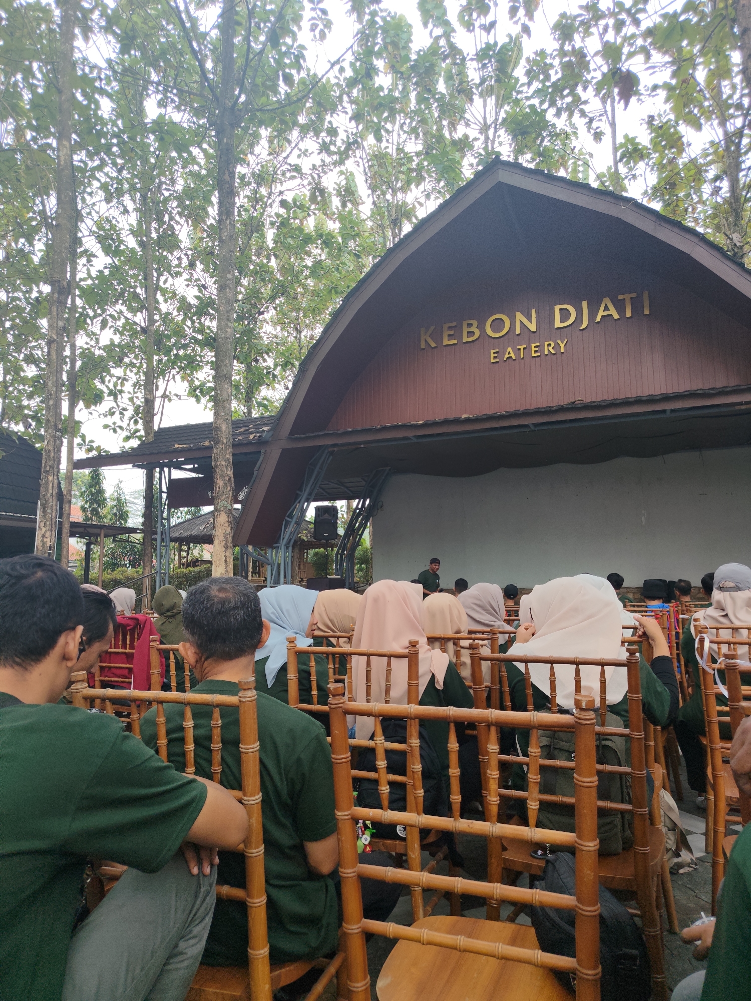 The image depicts an outdoor gathering at "KEBON DJATI EATERY" in Tasikmalaya, Indonesia. A group of people, many wearing green shirts and some wearing headscarves, are seated on rustic wooden chairs arranged in rows facing a stage area. The stage has a large, blank screen or backdrop and a roof structure above. Trees with green foliage surround the venue, suggesting a natural or park-like setting. The lighting indicates it is daytime, possibly late afternoon or early evening. The overall atmosphere appears to be one of an informal event or performance.