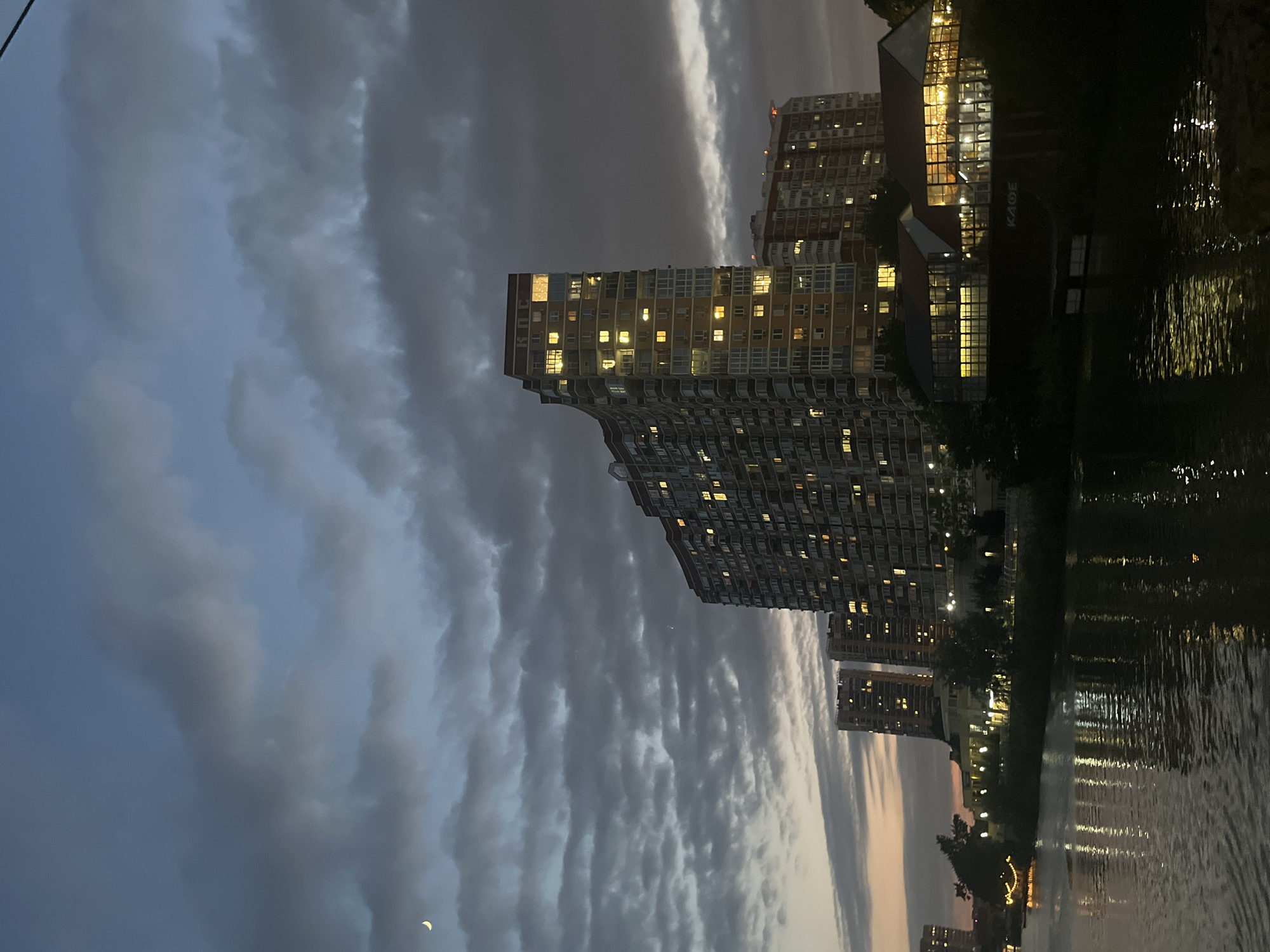 The image depicts a twilight scene in the city of Krasnodar, Russia, overlooking the Kuban River. In the foreground, the dark, rippling water of the river reflects the lights from the city. Across the river, a row of modern apartment buildings stands tall, their windows illuminated with a warm glow. The largest building, in the center, has a distinctive curved facade. To the right of the main apartment complex, a single-story building with large glass windows is visible, appearing to be a commercial establishment with a sign that reads "КАФЕ". Trees and smaller buildings line the riverbank between the water and the apartment complexes. The sky above is a dusky blue, filled with dramatic, dark clouds, with a sliver of a crescent moon visible in the upper left quadrant. The overall atmosphere is calm and serene, capturing the transition from day to night in an urban waterfront setting.
