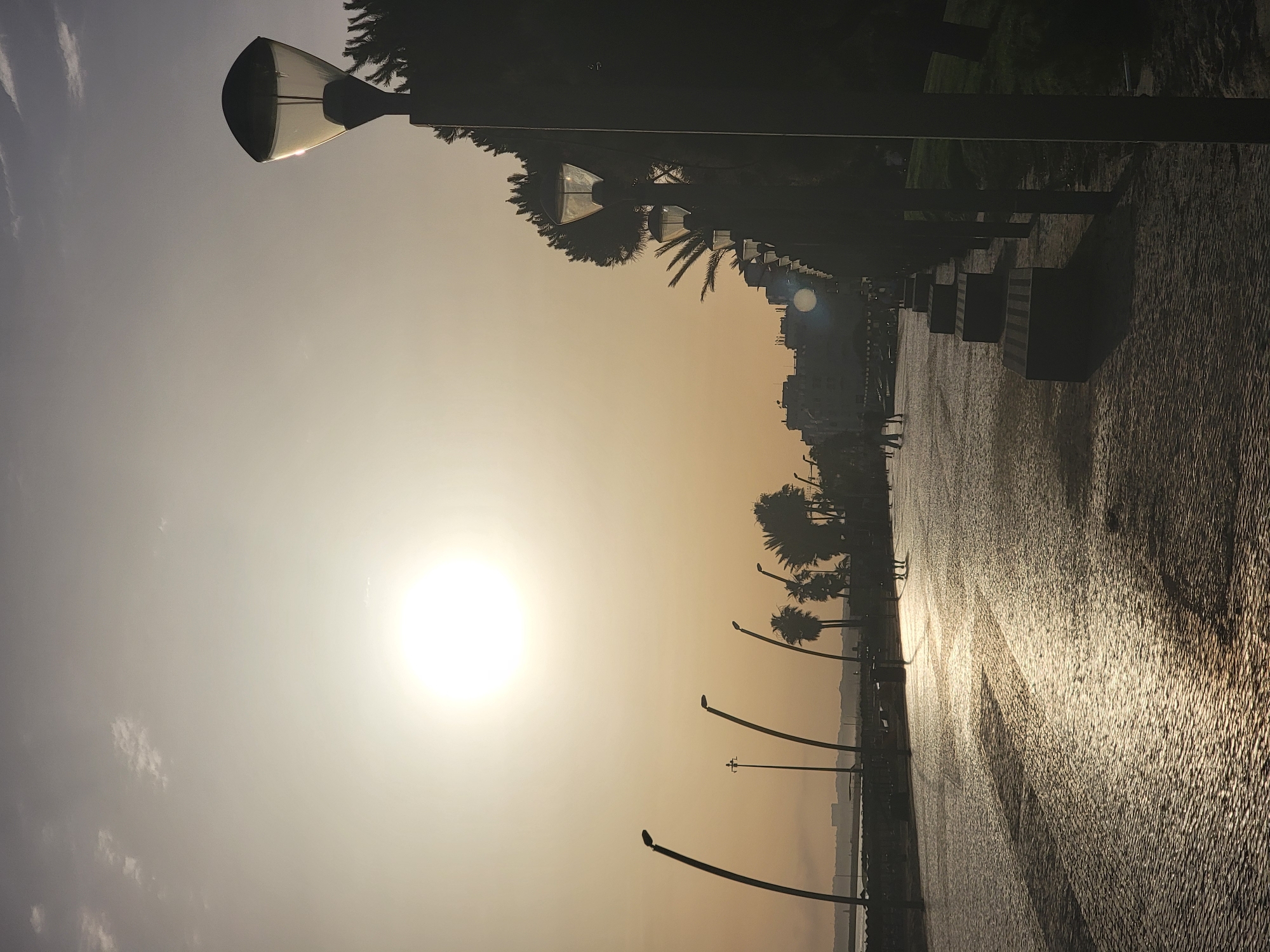 The image captures a sunset scene along the Quarteira promenade in Portugal. The low sun, positioned in the upper left quadrant, casts a warm, golden light that reflects off the wet cobblestone pavement of the walkway. Silhouettes of palm trees and other vegetation line the right side of the promenade, interspersed with street lamps and dark benches. In the distance, a faint outline of buildings suggests the urban setting. Several figures, also in silhouette, are seen walking along the promenade, their presence indicated by their indistinct forms. The overall atmosphere is serene, with the setting sun creating long shadows and a tranquil ambiance.