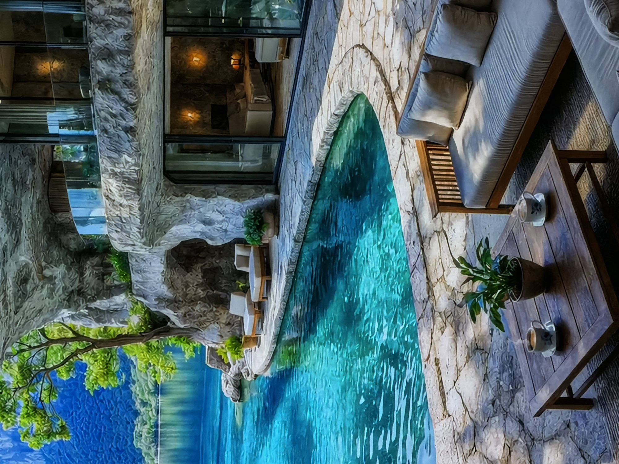 The image displays a luxurious architectural complex, possibly a resort or private residence, seamlessly integrated into a natural, rocky landscape. The multi-level structure is characterized by its robust stone cladding and extensive glass windows, which reflect the surroundings and reveal warmly lit interiors.
In the foreground, a sparkling turquoise swimming pool with gently rippling water parallels the building. The pool area is paved with light-toned stone, leading to an inviting outdoor lounge space on the right. This area features a modern wooden sofa with light-colored cushions and pillows, alongside a matching wooden coffee table. On the table, a potted green plant and two white mugs, one appearing to hold a beverage, are visible. Soft light, possibly from setting sun or artificial sources, casts gentle shadows across the furniture and stone work.
Below the complex, a vast expanse of calm, deep blue water stretches to the horizon, reflecting the sky and distant land. Lush green trees with dense foliage are prominent in the lower left, appearing to grow from the rocky terrain and overhang the water. No people are present, and there is no visible text. The overall scene conveys an atmosphere of tranquility, luxury, and harmonious coexistence with nature, likely set during late afternoon or early evening as indicated by the interior lighting and shadows.