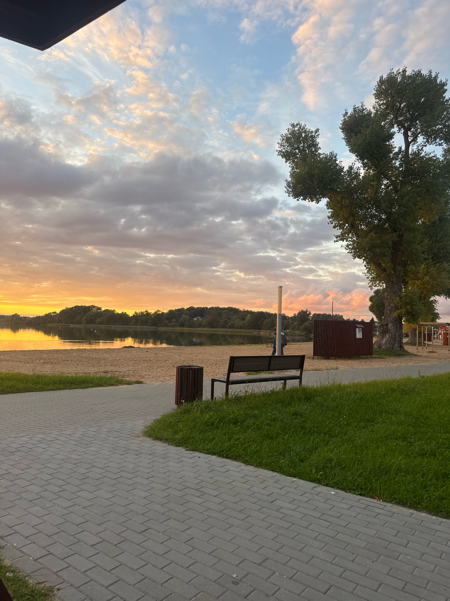 The image depicts a tranquil sunset scene at a lakeside beach in Myadzyel, Belarus. A paved walkway with grass borders leads towards a sandy beach bordering a calm lake. The sky is filled with a mixture of soft pink and orange clouds, indicative of sunset, contrasting with patches of blue sky and darker clouds.
Dominating the right side of the frame is a large, mature tree with lush green foliage. On the beach, a dark-colored bench faces the water, with a wooden trash receptacle nearby. In the background, several structures are visible, including a small, dark wooden building and some lighter-colored structures that might be part of a playground or recreational area. A tall, slender pole stands near the wooden building.
Further out on the lake, a tree-lined shore stretches across the horizon. A few distant figures can be seen on the beach in the mid-ground. The overall atmosphere is peaceful and serene, capturing the beauty of a natural landscape at dusk. The foreground is dominated by the textured gray paving stones of the walkway and a patch of vibrant green grass.