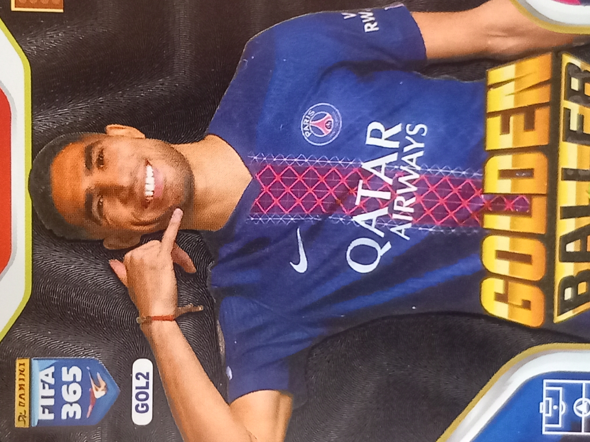 The image shows a close-up, medium shot of a male soccer player in a blue jersey. He is smiling and making a "call me" gesture with his right hand. The jersey has "QATAR AIRWAYS" written across the chest, and the Paris Saint-Germain crest is visible on the left side of the chest. The background is dark and textured. There are some text overlays, including "FIFA 365" and "GOL2" on the left side, and "GOLDEN BALLER" at the bottom. The lighting is bright, highlighting the player's face and jersey. The photo appears to be a collectible card or poster. There is no identifiable city or specific location visible in the image.