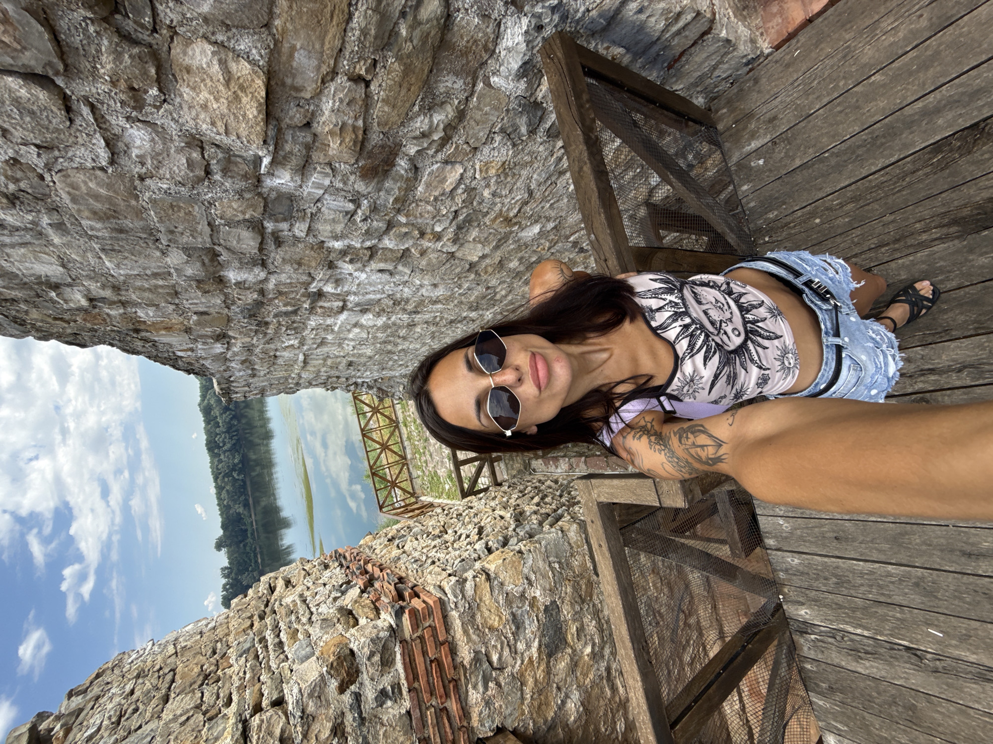 A woman with dark hair and sunglasses takes a selfie on wooden stairs within the ruins of a stone fortress. She is wearing a crop top with a sun design and denim shorts. The stairs lead down to a view of the Danube River and a treeline under a cloudy blue sky. The stone walls of the fortress are visible on either side of the stairs.