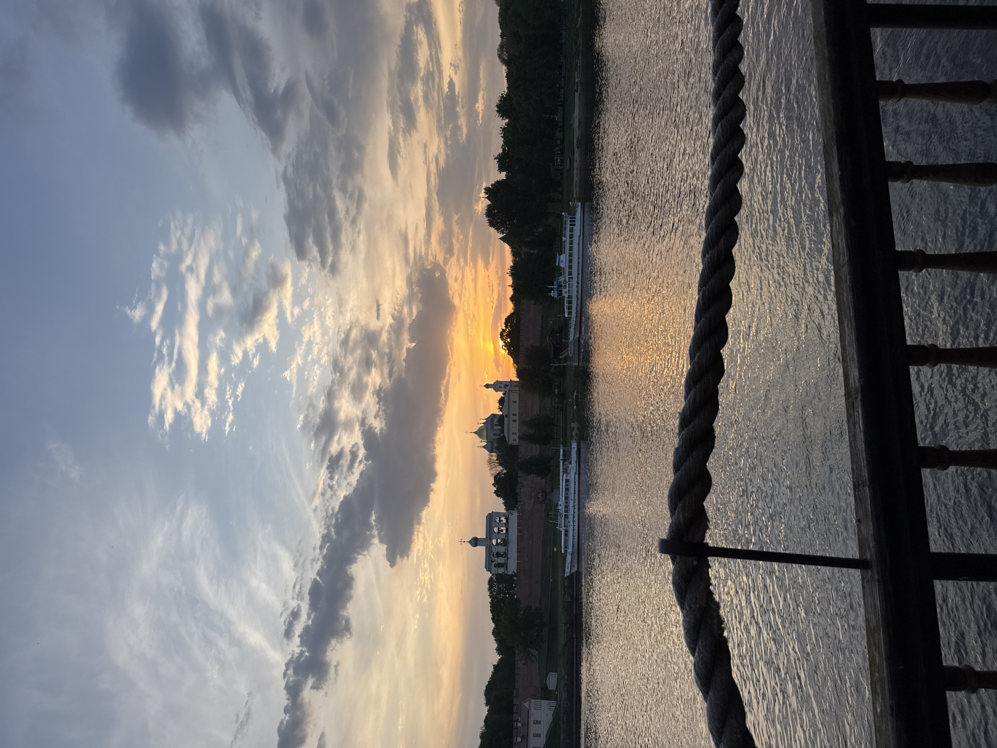 A vertically oriented image captures a sunset scene in Veliky Novgorod, Russia, viewed from a boat or pier. To the left, the sky transitions from a light blue to orange near the horizon, displaying scattered clouds. The setting sun casts a golden glow over the water. Across the water, the cityscape includes the Kremlin, visible with its distinctive white-walled structures and golden domes of cathedrals. A white riverboat is docked along the embankment. In the foreground, a thick rope runs vertically along the right edge, attached to a dark metal railing, partially obscuring the view of the water and the city. The water reflects the sunlight, creating bright streaks. The image suggests a tranquil evening setting.