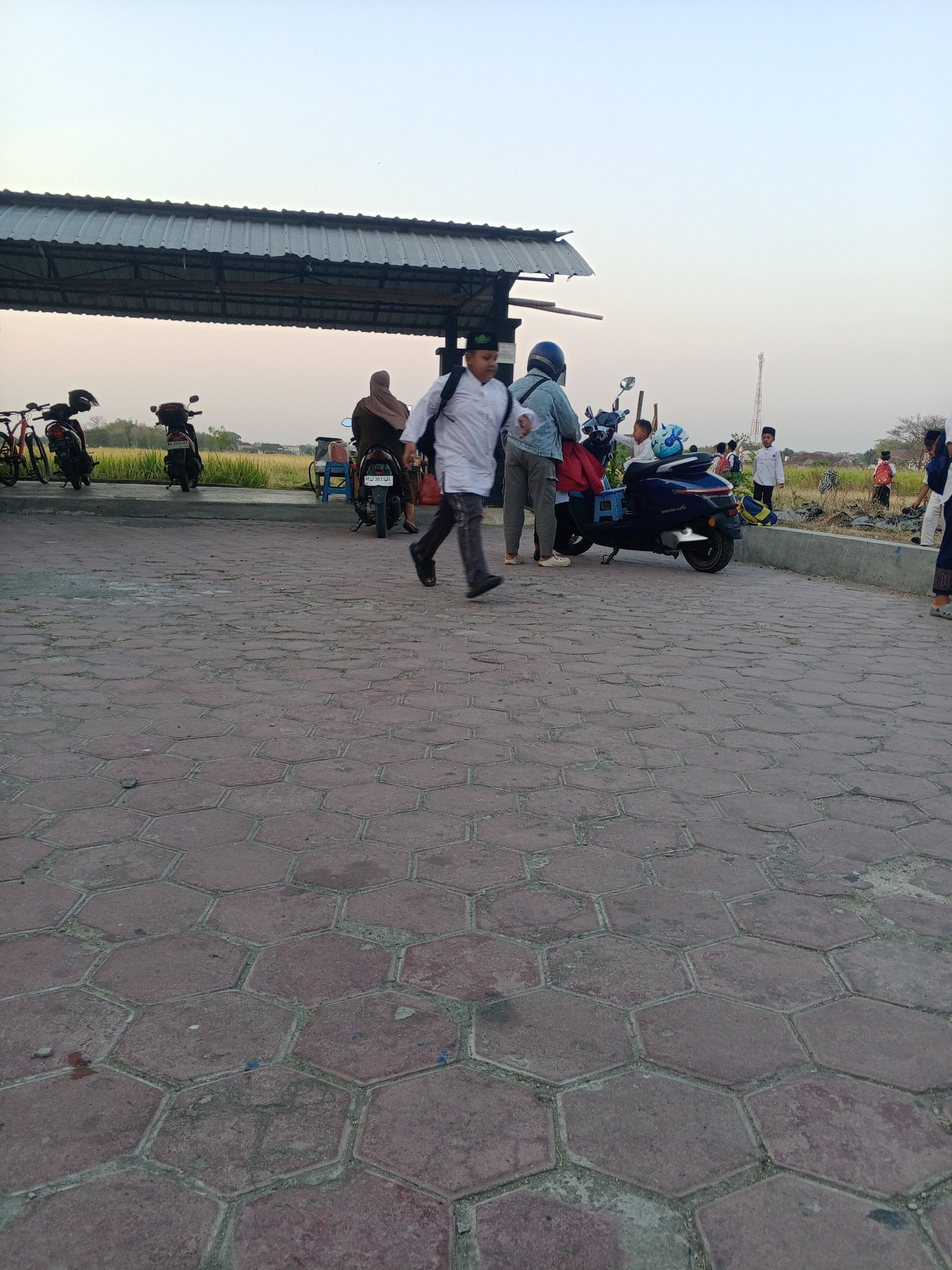 In Lembah, Indonesia, a group of people are gathered near a covered structure, possibly a bus stop or marketplace. The ground is paved with hexagonal red tiles. Several motorcycles are parked, one of which is blue and being approached by a person in a denim jacket. A young boy in a white shirt and cap is also near the blue motorcycle. A man in a white shirt and dark pants is walking past the scene. In the background, there are fields, trees, and a communication tower. Other individuals, including children, are visible in the distance, some with backpacks. The sky is clear and pale, suggesting it is either early morning or late afternoon.