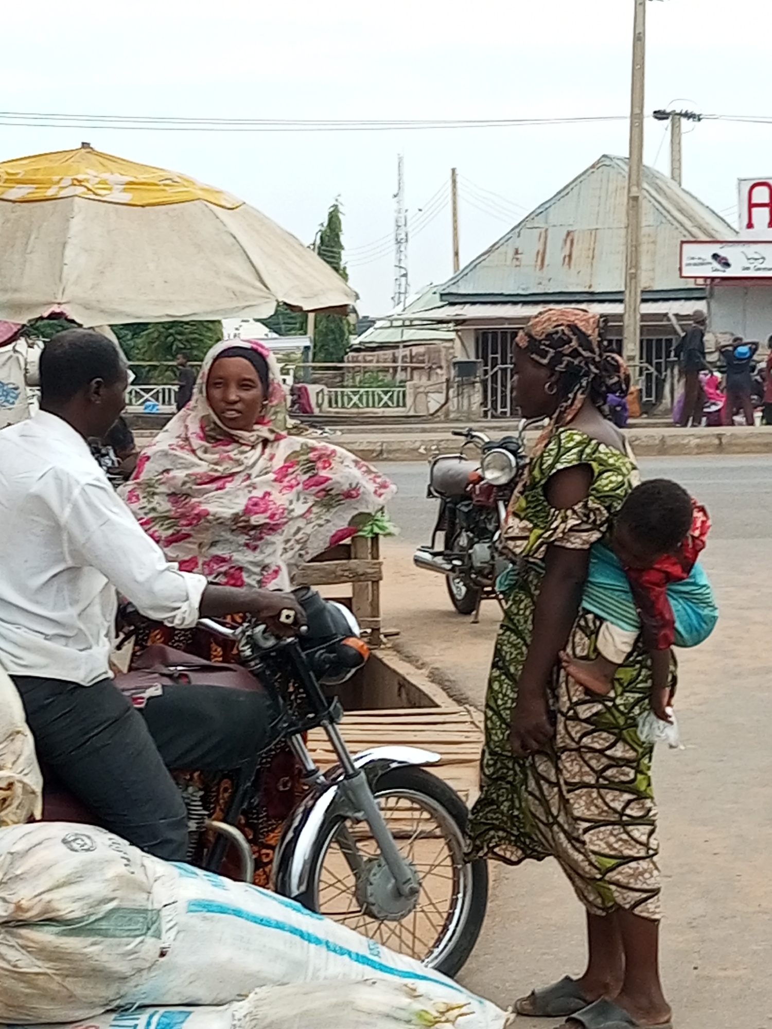 In Gombe, Nigeria, a man in a white shirt is seated on a motorcycle, facing a woman who is also on the motorcycle. The woman wears a floral headscarf and a matching patterned dress. To their right, another woman stands, carrying a baby on her back, secured with a colorful wrap. The baby is dressed in red and blue. The scene appears to be an outdoor market or roadside, with a yellow and white umbrella visible behind the man and woman on the motorcycle. Bags of goods are stacked in the foreground. In the background, buildings with corrugated iron roofs and a street with other people are visible. A power pole and overhead wires are also present. The overall atmosphere suggests a bustling urban environment.