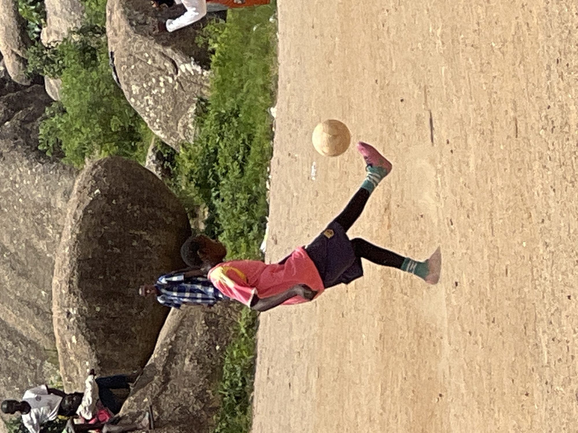 A young boy in a pink shirt and dark shorts is kicking a light brown soccer ball on a dirt field in Jos, Nigeria. He is wearing colorful socks and pink shoes. In the background, several large rocks and green bushes are visible. Other people are scattered around the scene, some sitting on the rocks. The sun is shining, casting shadows on the ground.