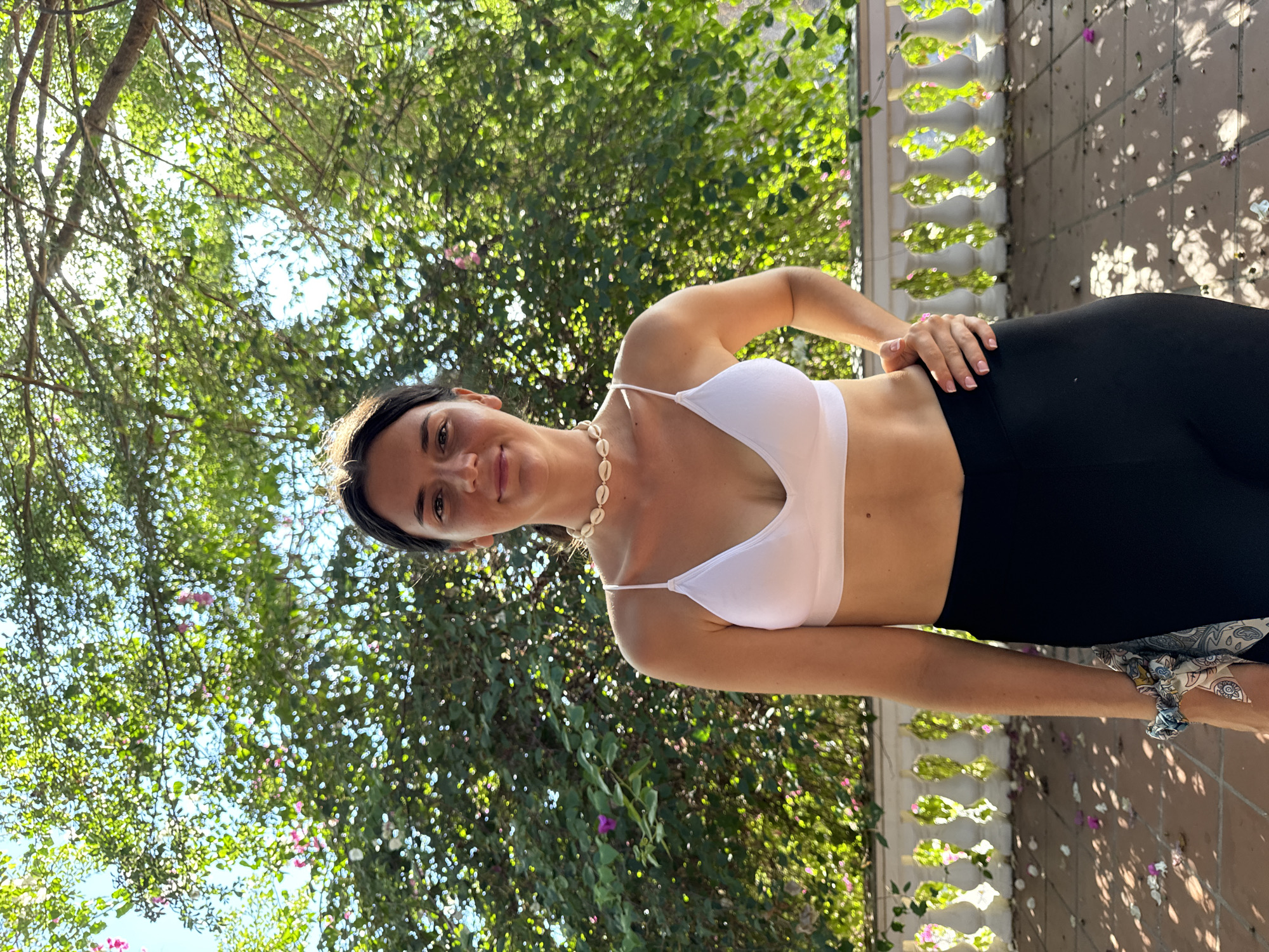 A young woman stands on a tiled surface in Mũi Né, Vietnam. She wears a white bra top and black leggings. She has a necklace and a scarf around her wrist. The background features lush green trees and a decorative white railing. Scattered flower petals lie on the ground. Sunlight filters through the leaves, casting shadows.