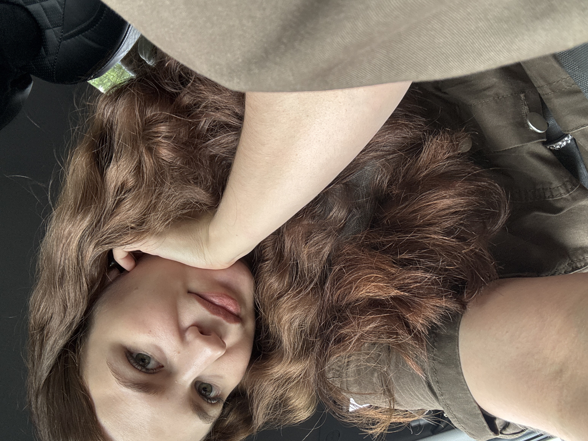 The image is a close-up, indoor selfie of a young woman with long, wavy brown hair. She is resting her head on her hand, with her elbow visible. She is wearing a brown collared shirt. The background is dark, with a hint of a textured black object, possibly a car seat, on the right side of the frame. There is also a sliver of green visible in the upper right corner, suggesting the presence of foliage outside. The woman is looking towards the camera with a slight smile. The overall impression is a casual, personal photograph. There are no discernible landmarks or features that would place this image specifically in the city of Chaikovsky, Russia.