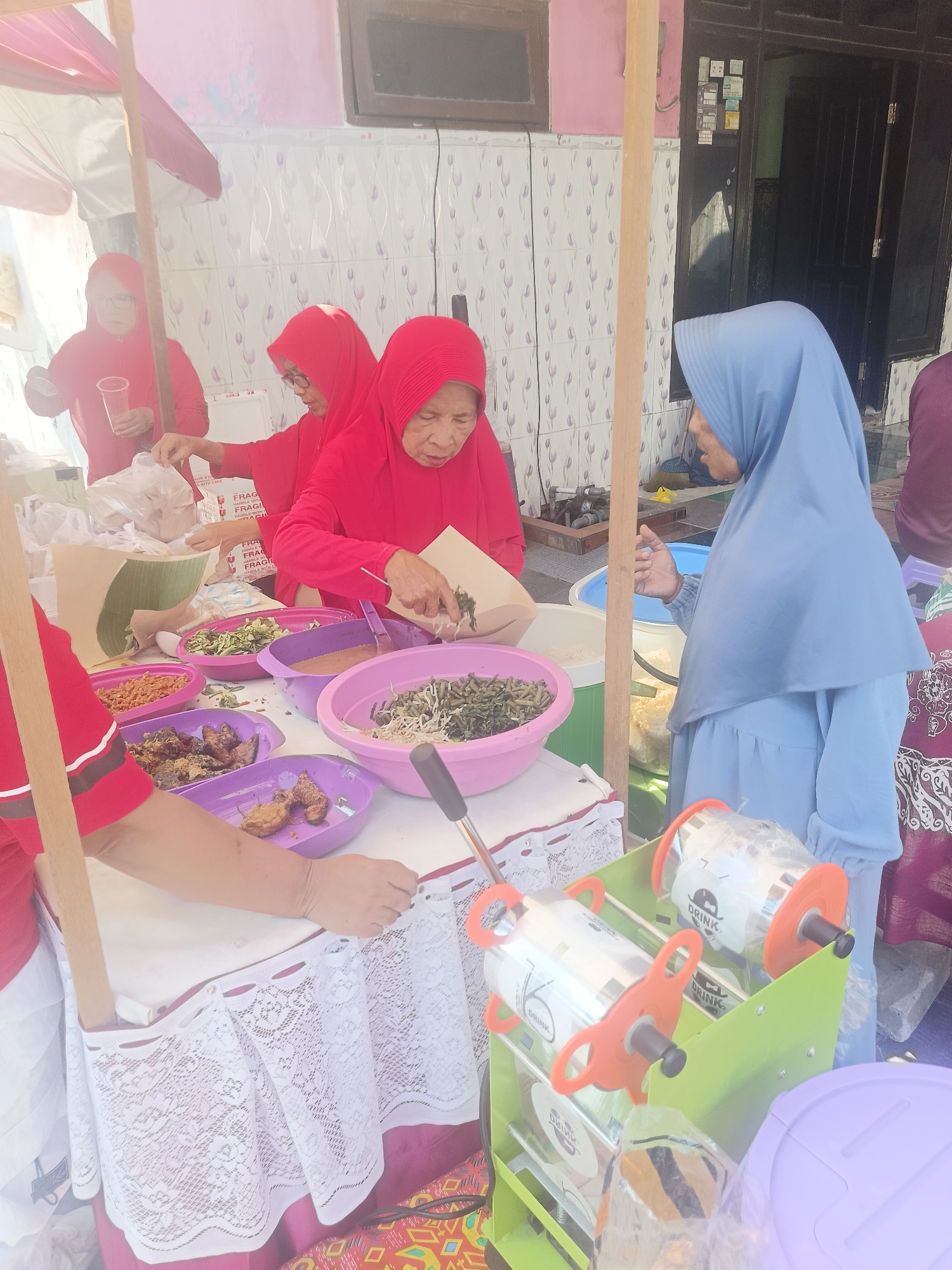 An outdoor food stall is set up in what appears to be a residential area in Gedangan, Indonesia. The stall has a white table covered with a white lace tablecloth. On the table are several bowls of food, including what looks like fried snacks in purple bowls, green vegetables in pink and purple bowls, and a bowl of what appears to be peanut sauce. A woman wearing a red hijab and red top is serving food from a paper bag into a bowl, and another woman in a red hijab and red top is standing behind the table with a plastic cup. To the right, a young woman in a blue hijab and blue dress is standing near a beverage sealing machine with two rolls of plastic film. In the background, a tiled wall and a dark-colored door are visible. The scene suggests a community event or a local market activity.