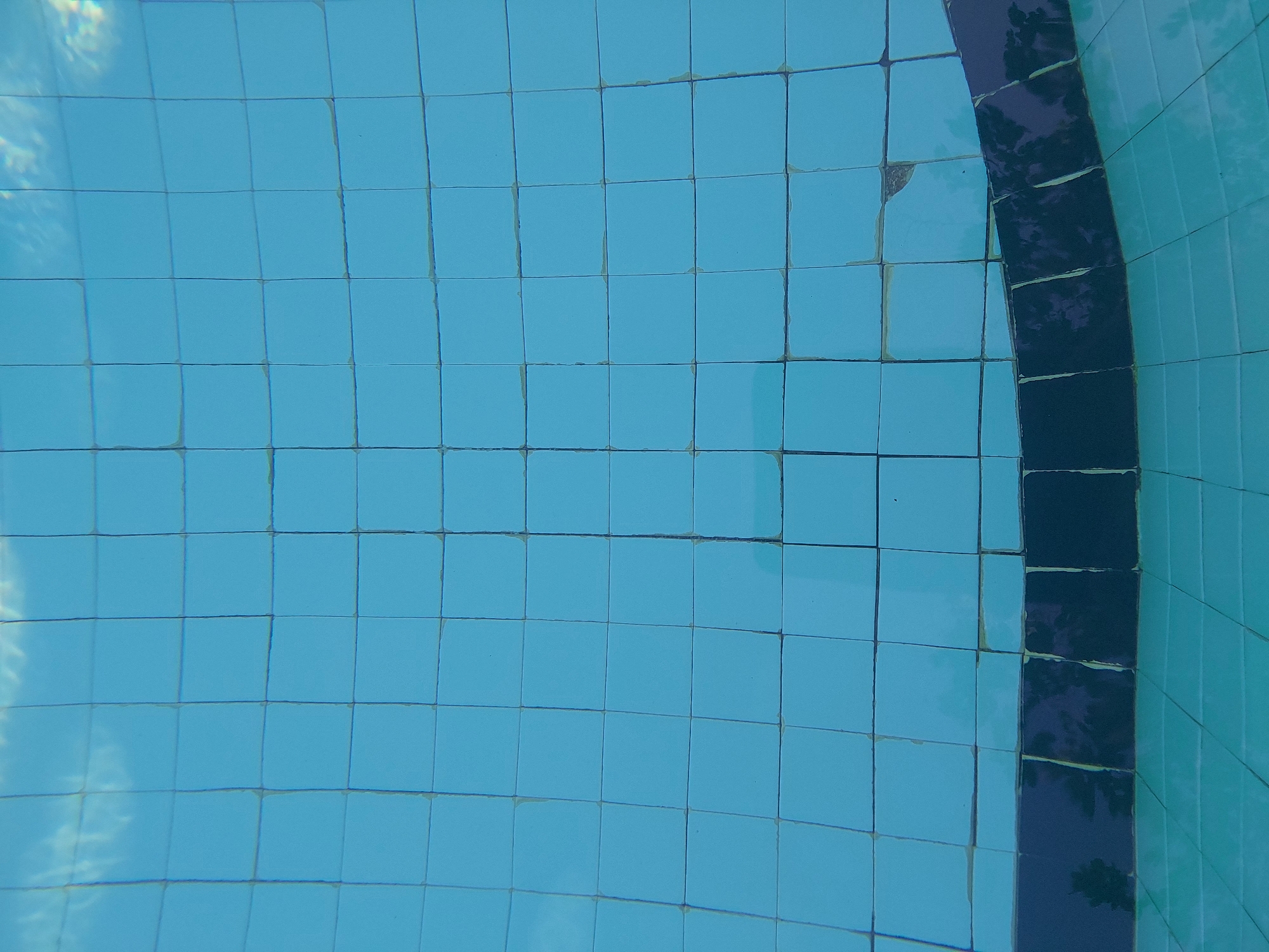 The image depicts an underwater view of a swimming pool in Baan Mae Nam, Thailand. The pool floor is covered in light blue square tiles, some of which show signs of wear and tear, with chipped edges and discolored grout. A darker blue tiled border curves across the bottom of the frame, reflecting what appears to be tree silhouettes. The water is clear, with subtle distortions and reflections of light and clouds visible on the surface. The overall scene is serene and captures the typical aesthetics of a residential or resort swimming pool.