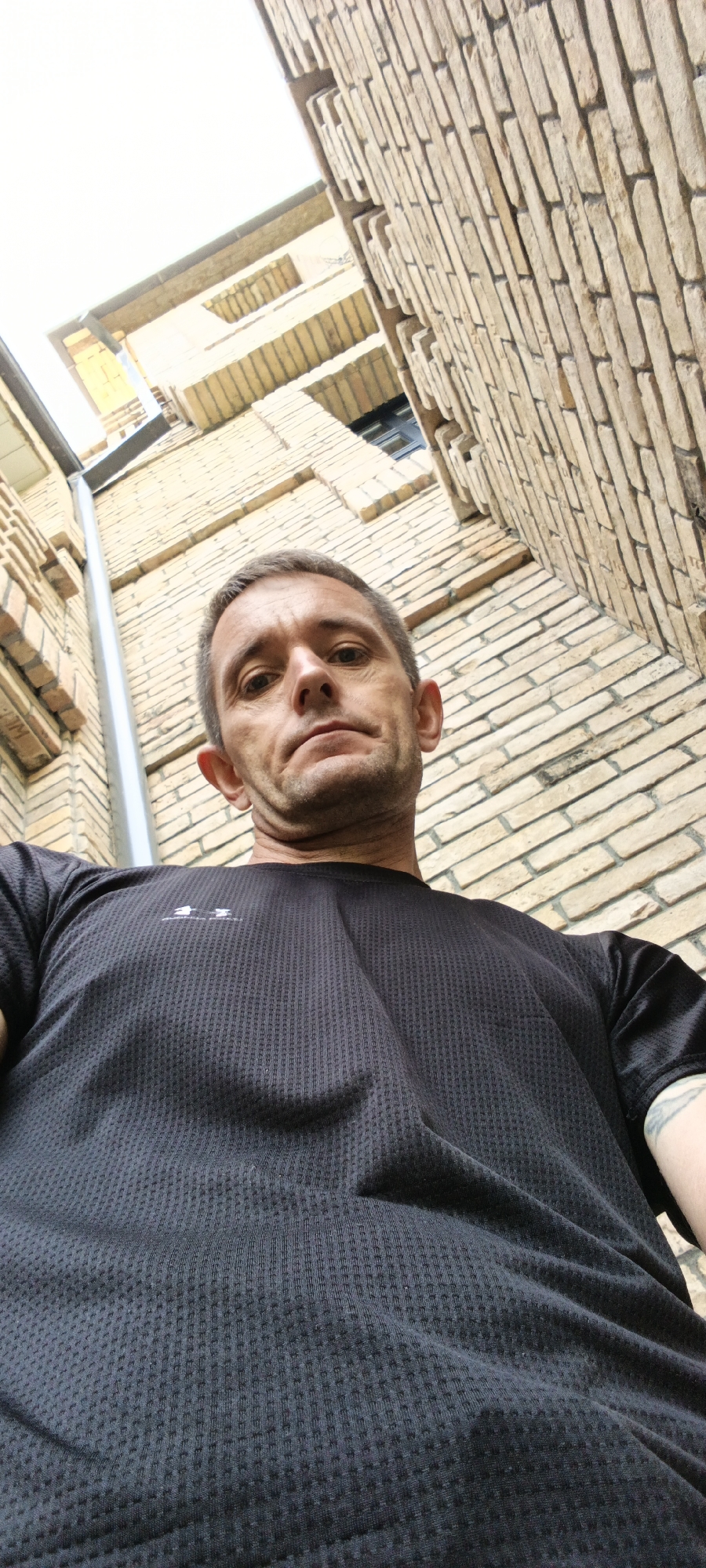 A low-angle, close-up, waist-up shot shows a man from the chest up, looking directly at the camera. He has short, graying hair and a fair complexion. He is wearing a black t-shirt with a textured fabric. Behind him, the image shows a yellow brick wall and a portion of a building with visible windows. The lighting suggests an outdoor daytime setting. The perspective implies the photographer is looking up at the man. The scene appears to be in an urban environment, possibly a courtyard or alleyway between buildings, consistent with urban architecture found in Tashkent, Uzbekistan.