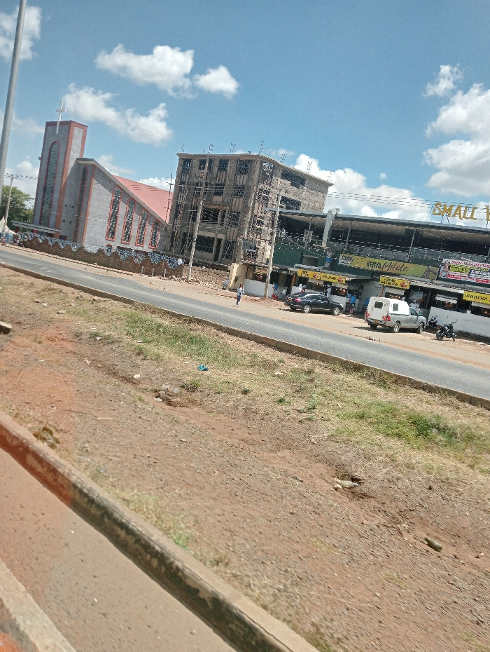 A street view shows a church with a tall steeple and cross to the left, adjacent to a building under construction. Further down the street is a shopping area with multiple stores. Cars and a motorcycle are parked along the roadside. A brown, grassy shoulder runs along the road, separating it from the vantage point. The sky is partly cloudy.