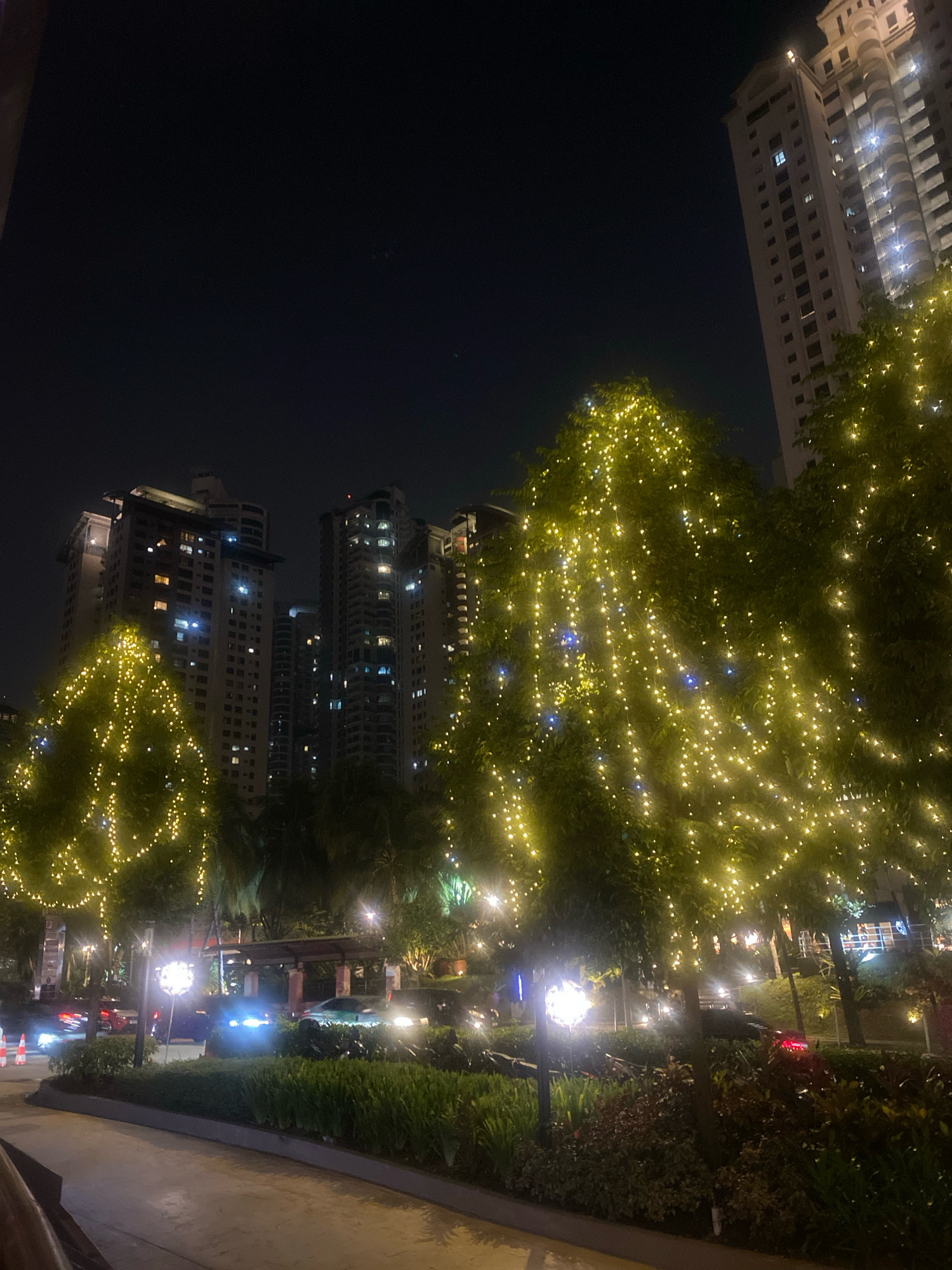 This is a night-time, street-level photograph taken in Kuala Lumpur, Malaysia, likely in a commercial or residential district. The foreground features a paved walkway and a manicured garden with various plants and some decorative lighting. Beyond the garden, a road is visible with several vehicles, some of which have their headlights on, illuminating the scene. Several trees are prominently featured throughout the image, adorned with strings of warm-toned fairy lights, creating a festive and illuminated atmosphere. In the background, tall residential or office buildings rise into the dark night sky. The buildings are illuminated by internal lights, with some windows visible. The overall impression is of a well-lit, urban environment at night, possibly a plaza or a street lined with apartments and trees decorated for an occasion. There are no visible people in the image.