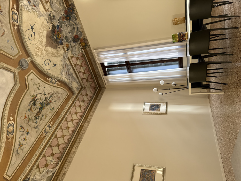 An interior view of a room, likely a palazzo, in Venice, Italy, is depicted. The primary feature is an elaborately frescoed ceiling with a central vaulted panel surrounded by segmented panels. The frescoes feature a mix of mythological or allegorical figures, swirling floral motifs, birds, and decorative borders in a warm palette of browns, creams, blues, greens, and pinks. A patterned frieze in pink and cream with a geometric floral design runs along the top of the walls.
The walls are painted a pale cream or off-white. On the left wall, two framed artworks with gold-colored frames and intricate, patterned designs are mounted. A vertical stripe of natural sunlight illuminates this wall between the two artworks.
To the right, a tall, dark-framed window with multiple panes is partially obscured by sheer white curtains, allowing natural light into the room.
The floor is a light-colored terrazzo-style finish with small, speckled aggregates. Part of a lighter, plain rug or mat is visible in the bottom left corner.
In the foreground, a modern dining or meeting area is furnished. A rectangular table with a white base and a dark, possibly black, tabletop is positioned with four dark gray or black chairs visible around it. To the left of the table, a tall, black multi-headed floor lamp with four spherical white diffusers stands. On the table, two decorative glass vases are present: one green and yellow with a coiled design, and another amber-toned with a textured surface. No individuals are present in the scene.