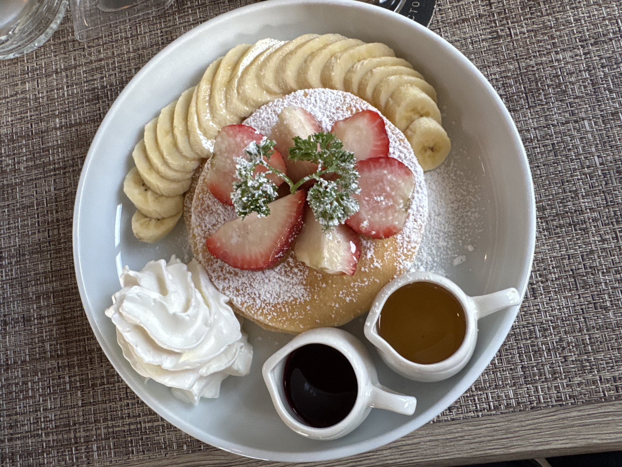 An overhead close-up shot captures a light blue rimmed plate holding a stack of pancakes adorned with sliced strawberries, powdered sugar, and a sprig of green garnish. To the right of the pancakes, a crescent of sliced bananas is arranged. The plate also contains two small white pitchers; one filled with a dark syrup and the other with a golden honey. A dollop of whipped cream sits on the left side of the plate. The plate is set on a woven placemat with a brown and tan textured pattern, and a wooden table is visible to the left and bottom of the frame. A sliver of a clear glass is visible in the upper right corner.