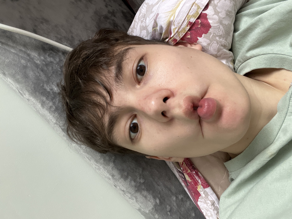 An individual is depicted in a close-up, front-facing selfie, taken indoors, presumably in Zainsk, Russia. The subject, with short brown hair and light skin, gazes directly into the camera with wide-set brown eyes. Their lips are intentionally pursed and pushed outwards, forming a circular protrusion. The individual wears a light green crew-neck garment, with a thin silver chain visible at the base of the neck. The background consists of a grey, textured, possibly upholstered surface on the left and a pillow with a patterned pillowcase, featuring red and maroon floral elements on a light ground, on the right. A portion of a light-colored wall is visible in the upper left.