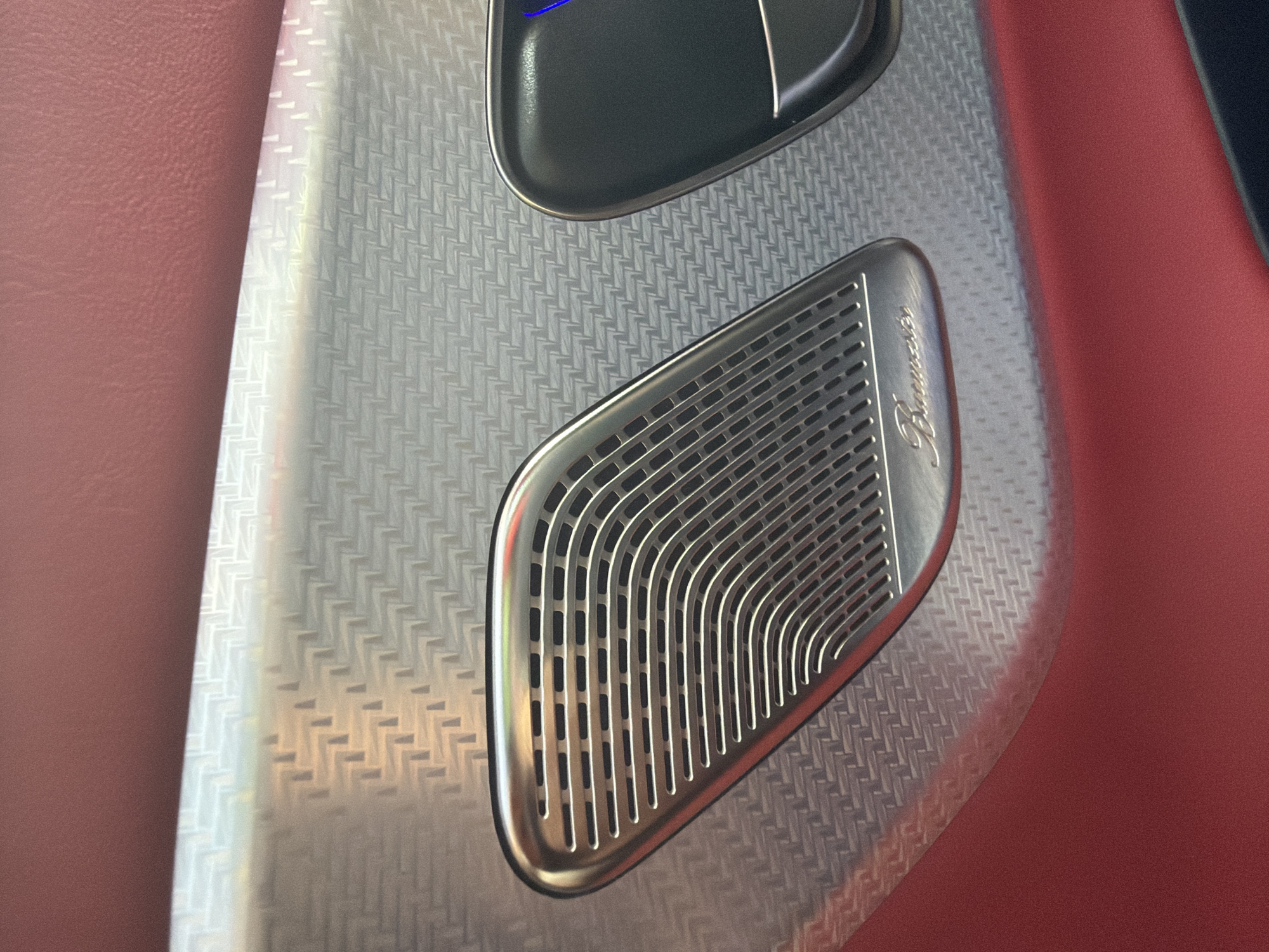 The media file presents a close-up view of a luxury car interior, highlighting intricate details of the door panel. The primary focus is on a metallic speaker grille and a textured panel, flanked by red upholstery.
On the right side of the image, a sophisticated, silver-colored speaker grille is prominently featured. It has an elongated, somewhat irregular pentagonal shape with a smooth, polished bezel. The grille itself displays an elegant design of vertical and gently curved horizontal slots, creating a distinct pattern. Engraved in script on the upper right part of the grille is the word "Burmester," indicating the brand of the premium sound system.
The speaker grille is set into a wider panel that exhibits a fine, repeating herringbone or woven texture in a light silver or grey tone. This textured pattern creates a sense of depth and sophistication. Above the speaker and partially visible, there is another dark grey or black panel element, likely part of a door handle or control unit, also outlined with a metallic trim. A subtle hint of blue light is visible at the very top, possibly from an illuminated button or indicator.
To the left and right edges of the image, a rich, reddish-brown upholstery material, likely leather, is visible. This material has a subtle grain and adds a contrasting color and texture to the metallic and textured panel elements. The lighting appears to be artificial, casting soft reflections on the metallic surfaces. No activity is taking place, and the image captures the static aesthetic of the vehicle's interior. The specific location of Trastenik, Bulgaria, is provided as context but is not visually represented in this interior shot.