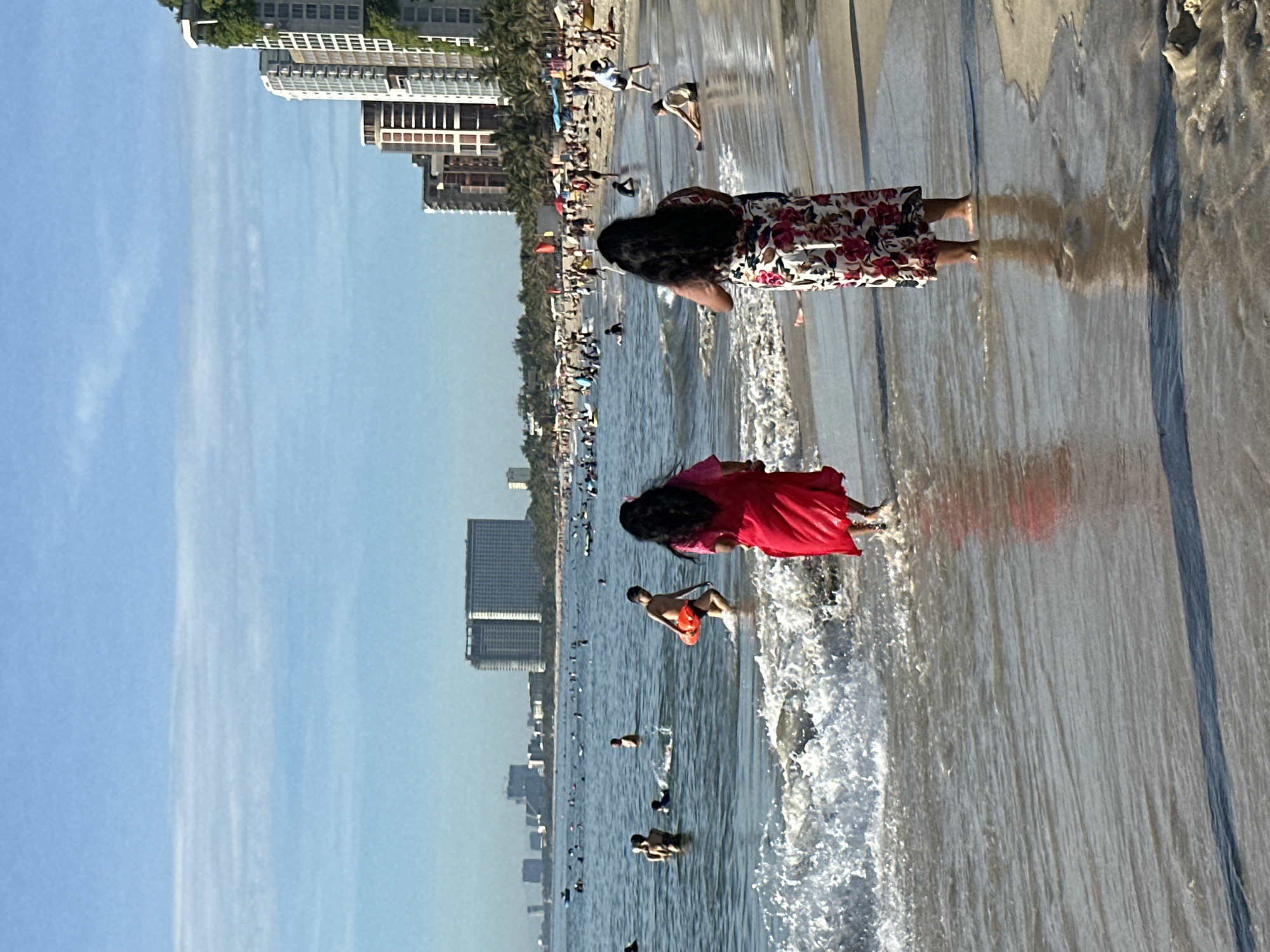 This image captures a bustling beach scene in Đà Nẵng, Vietnam, likely along My Khe Beach, known for its long stretch of sand and clear waters. In the foreground, two women are seen wading into the shallow ocean. The woman on the left wears a vibrant red dress, while the woman on the right is dressed in a white floral-patterned dress. Both are facing away from the camera, looking out towards the sea.
The water is calm with gentle waves lapping at the shore, creating white foam. In the middle ground, numerous people are enjoying the beach and water. Some are swimming, others are standing in the water, and a few are engaged in activities like playing or wading. A man in the water is holding an orange float.
The background is dominated by a cityscape with numerous tall buildings, characteristic of Đà Nẵng's modern development. These skyscrapers, some appearing residential or hotel complexes, line the coast, with palm trees interspersed among them. The sky is a clear blue with some thin, wispy clouds, suggesting a sunny day. The overall impression is one of a lively and popular tourist destination.