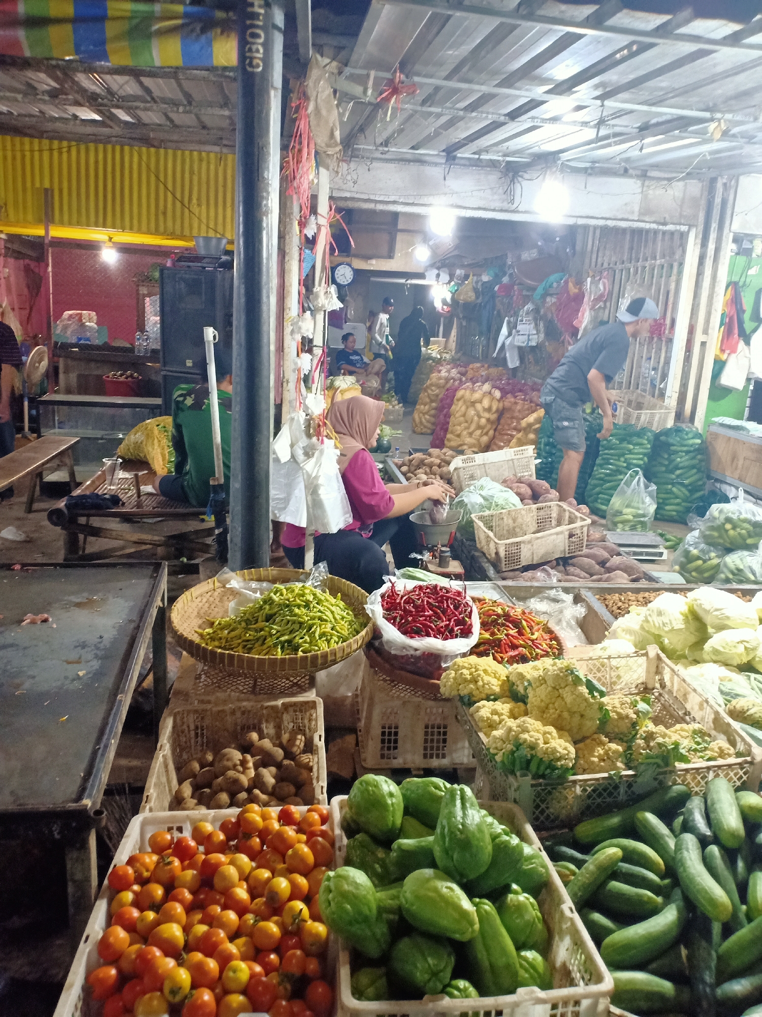This image captures a bustling scene at a traditional market in Semanding, Indonesia. The market appears to be an outdoor or semi-covered space, with a corrugated metal roof supported by metal poles and wooden beams. Various stalls are set up, displaying a colorful array of fresh produce. In the foreground, several large crates overflow with vibrant vegetables and fruits. One crate is filled with small, red tomatoes. Next to it, a crate holds numerous green, elongated gourds, possibly chayote. Adjacent to these, a bin is brimming with cucumbers. Further back, a woven basket holds a substantial pile of small green chilies. Another basket contains red chilies. Cabbages and cauliflowers are also visible in crates, alongside baskets of potatoes and what appear to be sweet potatoes.
In the middle ground, several individuals are engaged in market activities. A woman wearing a pink shirt and a headscarf is seated and appears to be weighing produce on a scale. Another person in a green camouflage shirt sits on a wooden bench, looking towards the scale. A man wearing a grey t-shirt and a cap is visible in the background, interacting with a pile of green bags, likely containing produce. Other market-goers and vendors are present, though their activities are less discernible. Bags of what seem to be onions or potatoes are stacked in the background. The overall atmosphere suggests a lively and active marketplace, typical of Indonesian local commerce.