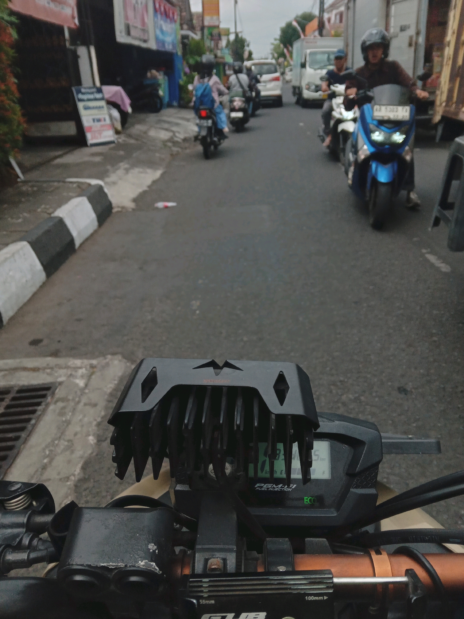 The image captures a street scene in Yogyakarta, Indonesia, from the perspective of a motorcycle rider. The foreground features the motorcycle's handlebars and instrument cluster, displaying "PGM-FI FUEL INJECTION" and an "ECO" indicator. A black, finned accessory is mounted above the instrument panel. The road ahead is visible, with other vehicles and pedestrians present. To the left, a sidewalk with black and white striped curbing lines the road, adjacent to buildings with signage. A person on a blue motorcycle is seen further down the street, as are several other motorcycles and a white truck. The environment appears to be a typical urban setting with moderate traffic.