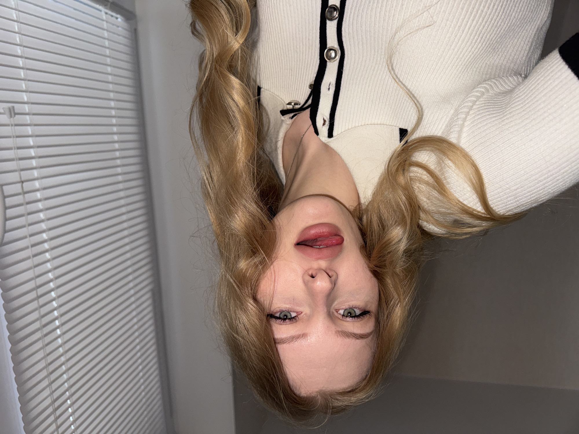 A young woman is indoors, posing for a selfie. She is fair-skinned, with blonde, wavy hair, and green eyes. She is wearing a white, ribbed cardigan with black trim. The background includes a white window blind. The location is unknown and there is no evidence that the image was taken in the city of Perm, Russia.