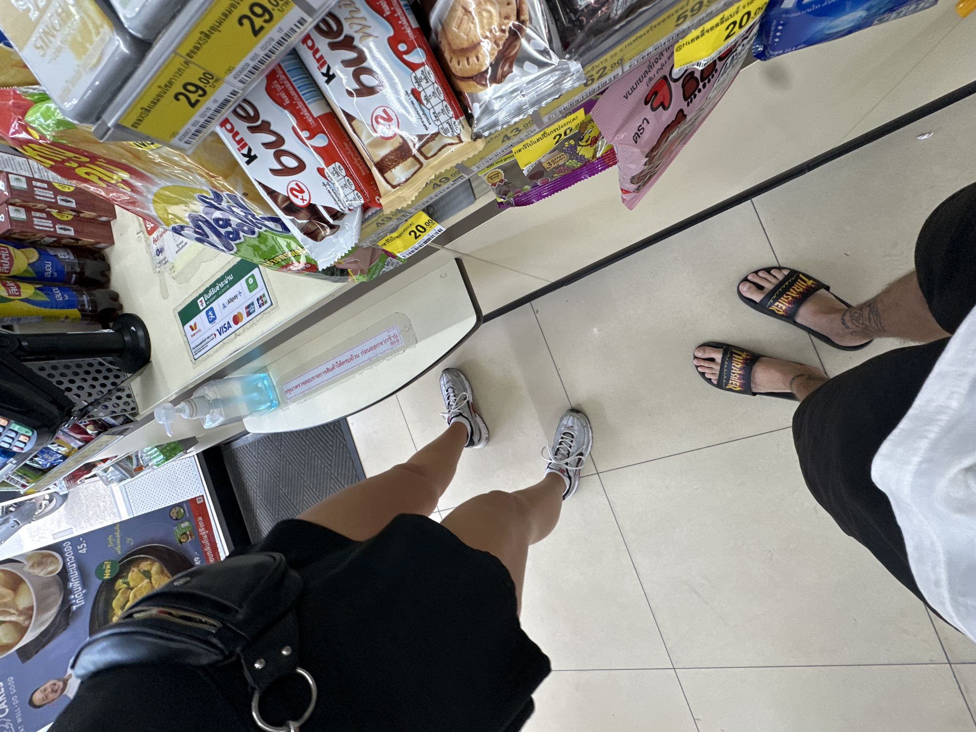 The image shows the feet of two individuals standing in what appears to be a convenience store in Karon, Thailand. The person on the left is wearing black shorts and black slide sandals with the word "THRASHER" in yellow and orange lettering. They have a tattoo on their left calf. The person on the right is wearing black shorts and silver athletic shoes with white laces and red accents. In the foreground, a variety of packaged snacks and candy bars are displayed, including several Kinder Bueno bars. There is also a bottle of hand sanitizer and a payment terminal visible on the counter. In the background, a menu board with food items and prices is displayed, and a person's head can be seen in the upper right corner of the image. The floor is tiled with a light-colored, rectangular pattern.